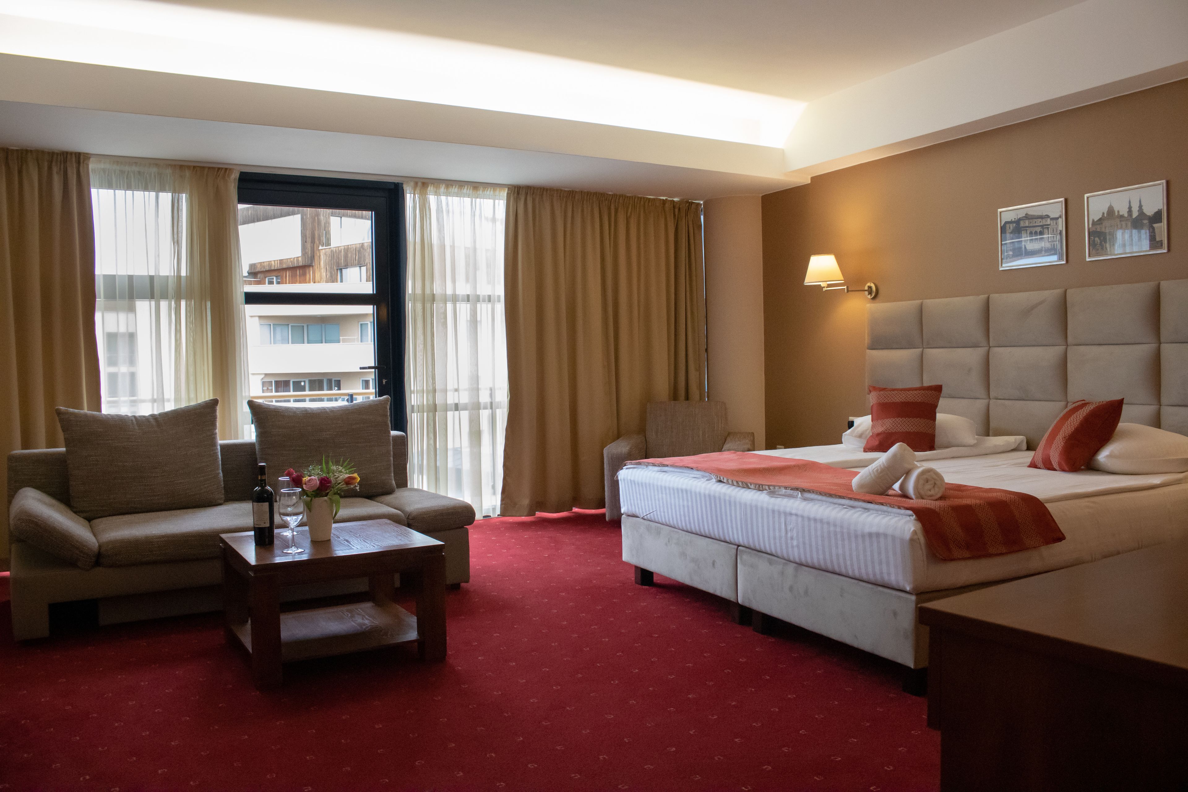 Una habitación de hotel con cama, sofá, una mesita con una botella y flores, y una ventana con cortinas.