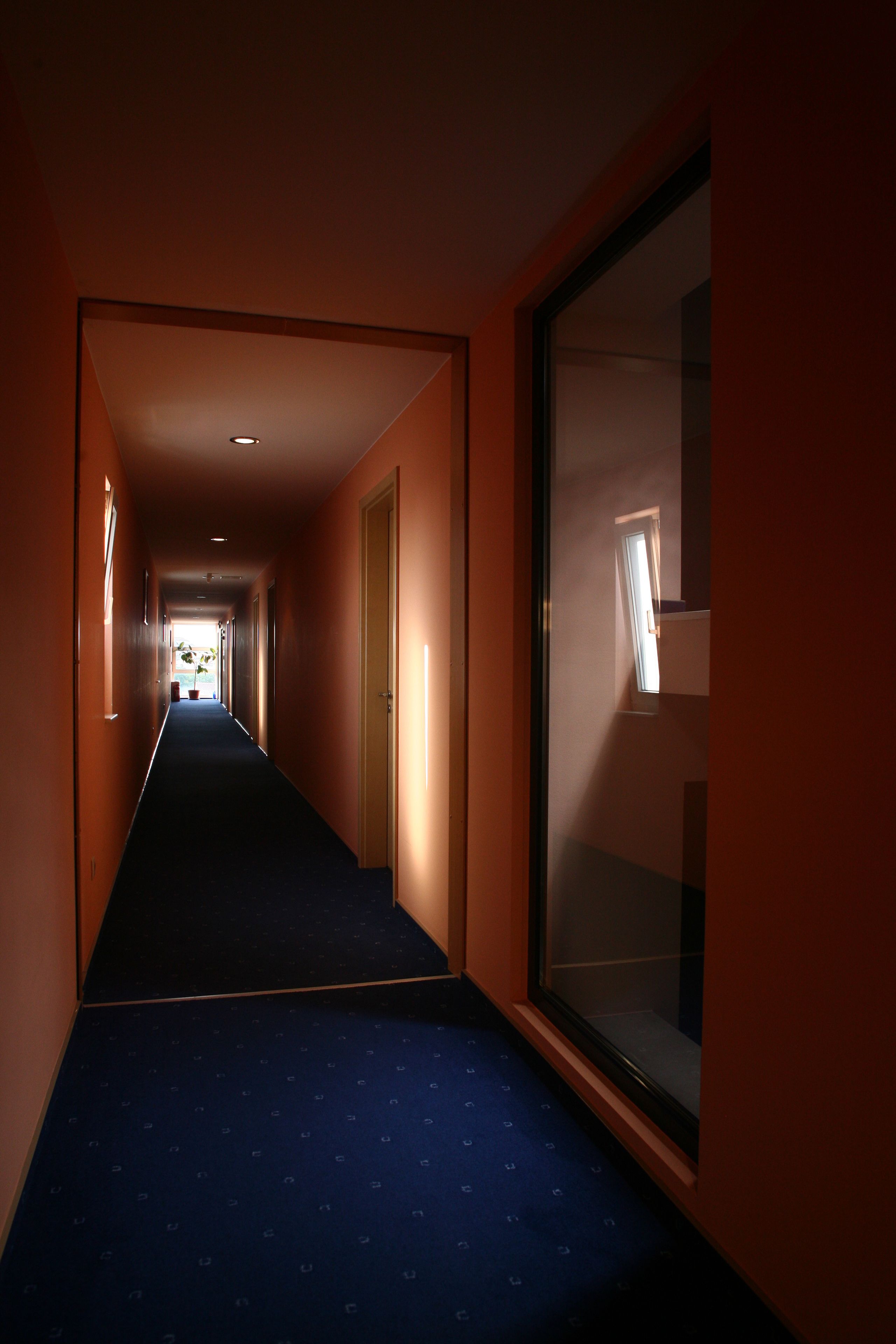A hotel corridor with a blue carpet, a large window on the right, and a door at the end.