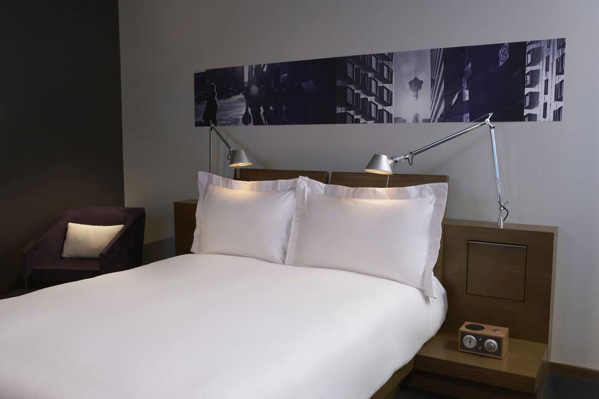 Quarto de hotel com uma cama, duas luminárias de cabeceira, uma cadeira, mesa de cabeceira e uma obra de arte pendurada na parede.