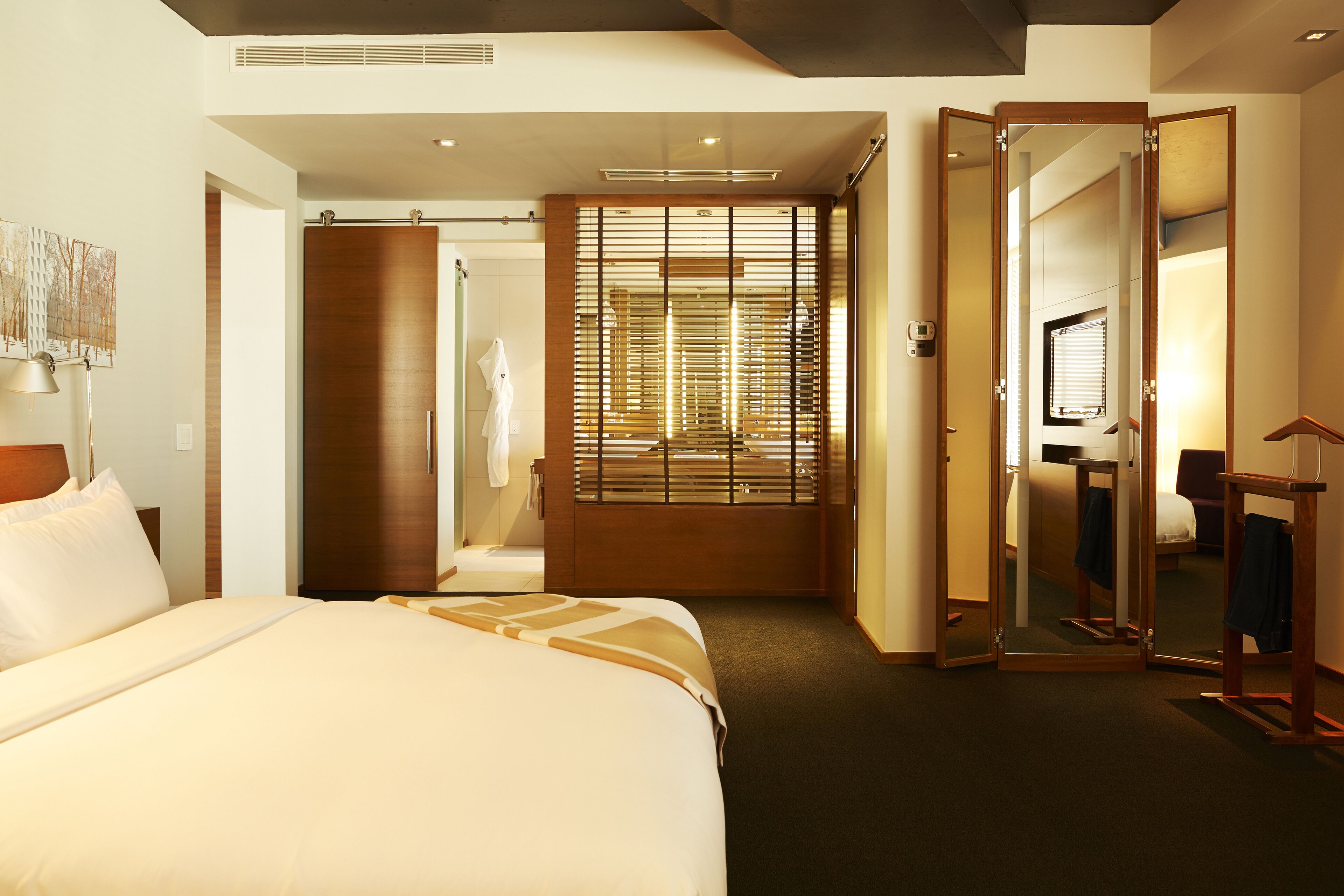 A hotel room with a large bed, a wooden wardrobe, a mirror, and a small table with a lamp.