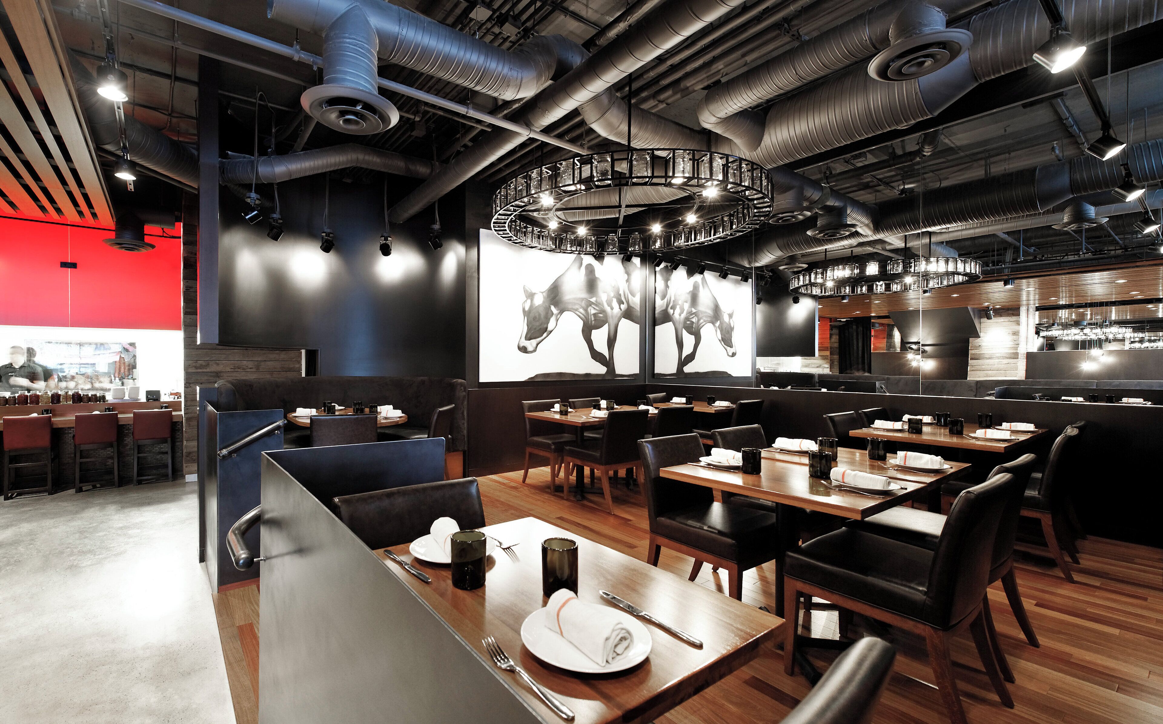 En modern restauranginteriör med högt i tak, synliga ventilationskanaler och en stor kristallkrona i mitten. Borden är dukade med vita tallrikar, silverbestick och svarta glas.
