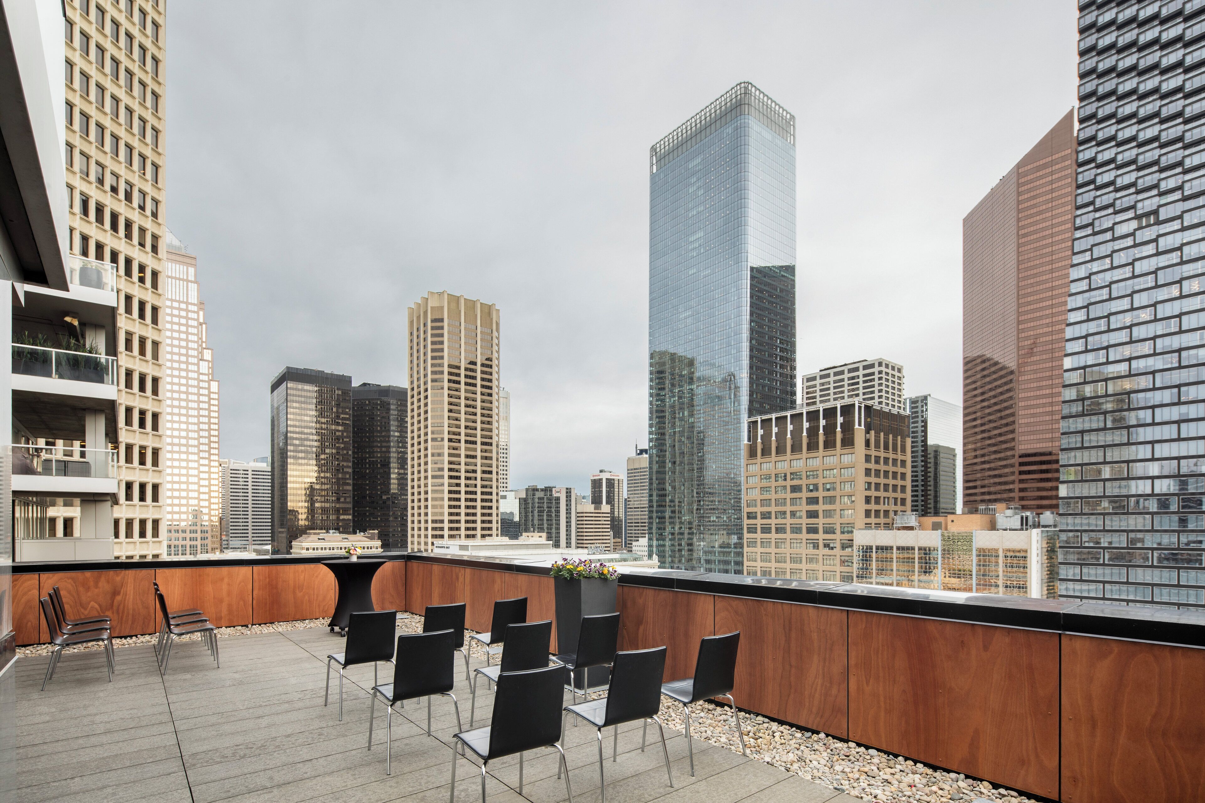 Terraza en la azotea con sillas modernas y una jardinera, con vistas a un paisaje urbano con rascacielos.