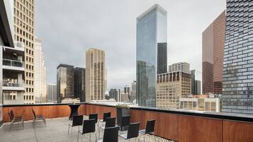 Terraza en la azotea con sillas modernas y una jardinera, con vistas a un paisaje urbano con rascacielos.