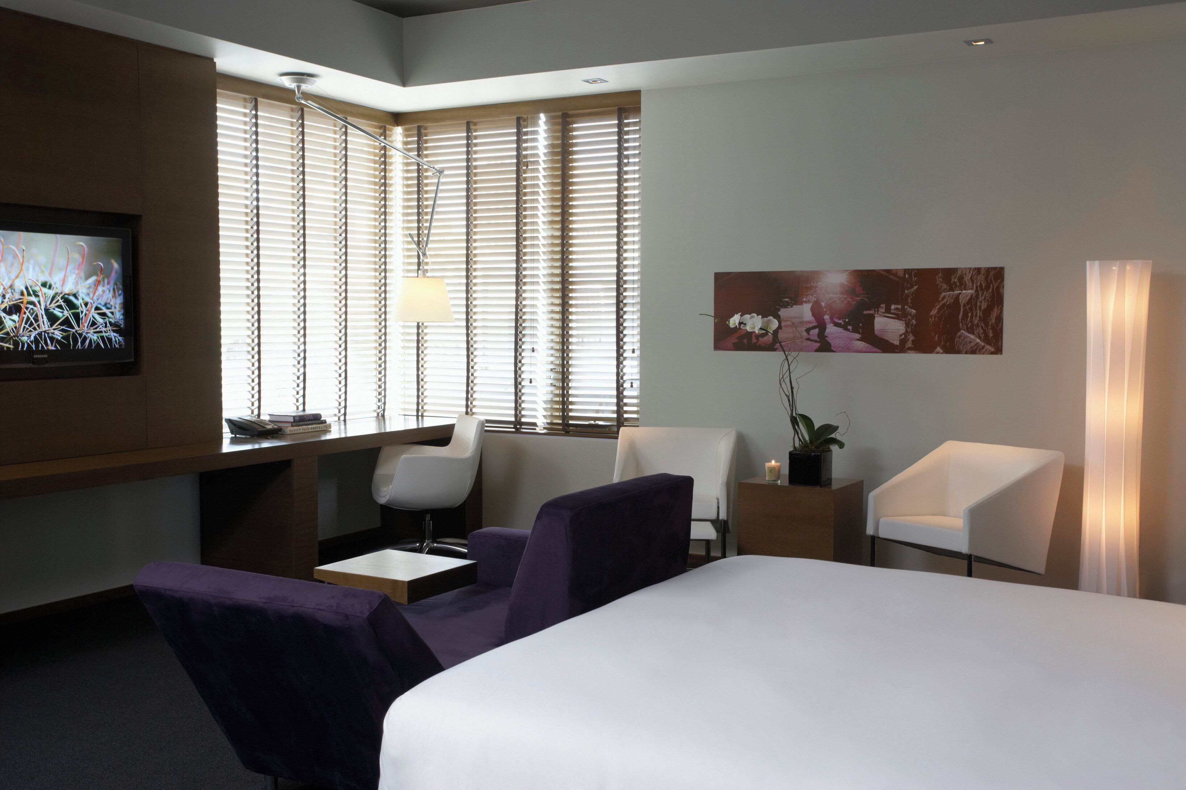 A hotel room with a bed, two chairs, a desk with a lamp, a TV, and a large window with blinds.