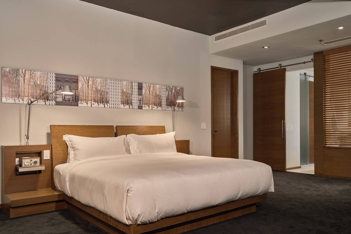 Quarto moderno com estrutura de cama de madeira, mesa de cabeceira com rádio e uma janela grande com persianas.