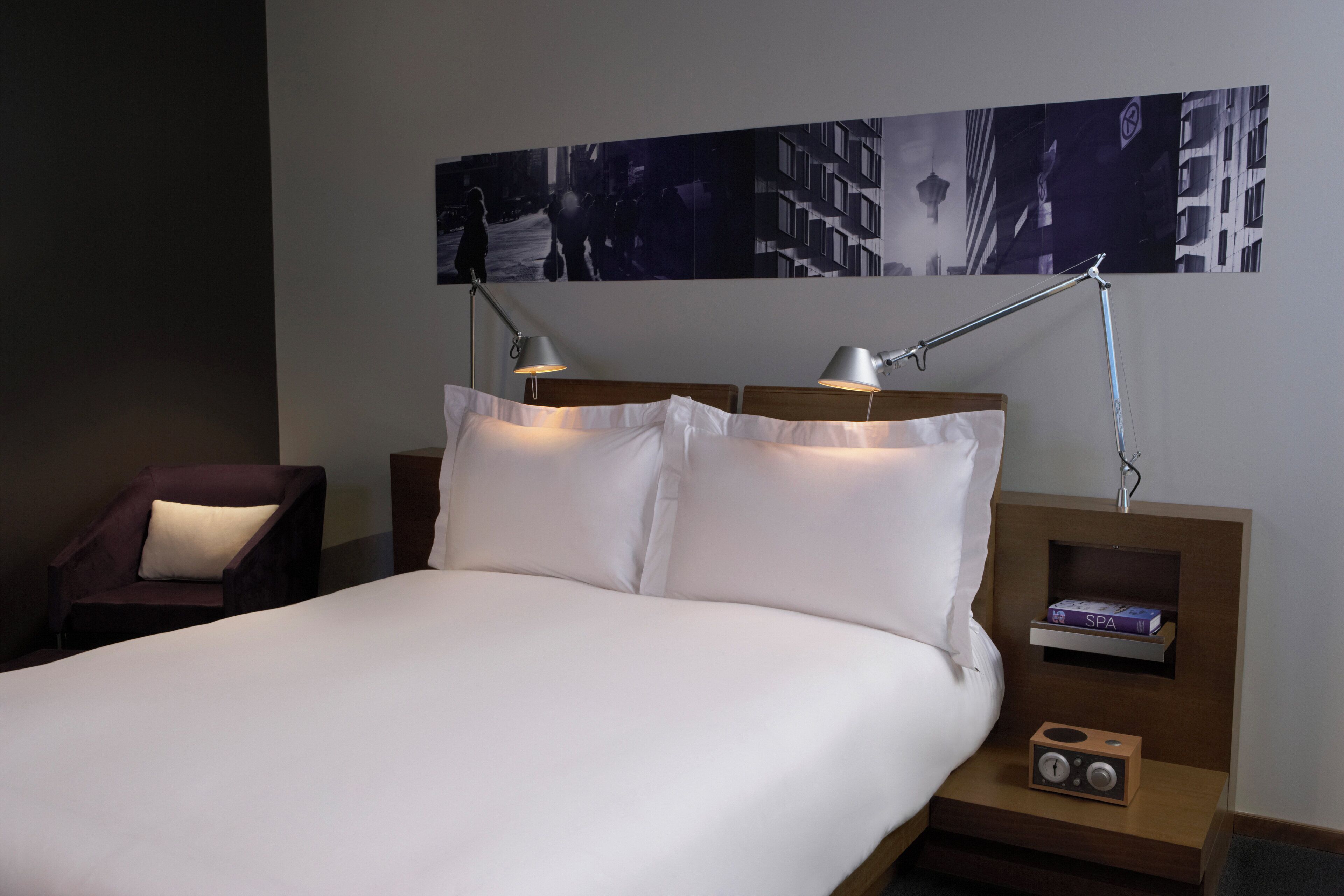 A neatly made bed with white linens, two bedside lamps, a small wooden nightstand with a box labeled "SPA", and a chair with a cushion.