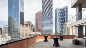 A rooftop terrace with a view of modern skyscrapers, featuring a wooden deck, two black tables, and potted plants. - Hotel Le Germain Calgary (Calgary)