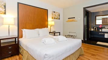Habitación de hotel con una cama grande, dos mesitas de noche, una mesilla, un cabecero de madera, un espejo, una pequeña mesa con lámpara y un baño con lavabo y toallero.