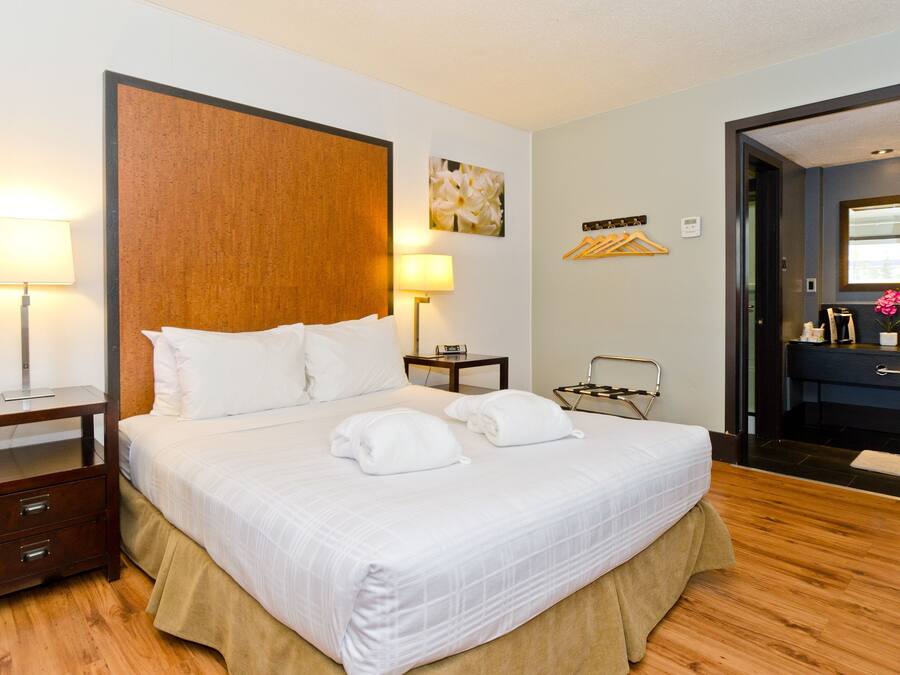 Une chambre d’hôtel avec un grand lit, deux tables de chevet, une commode, une tête de lit en bois, un miroir, une petite table avec une lampe, et une salle de bain avec un lavabo et un porte-serviettes.