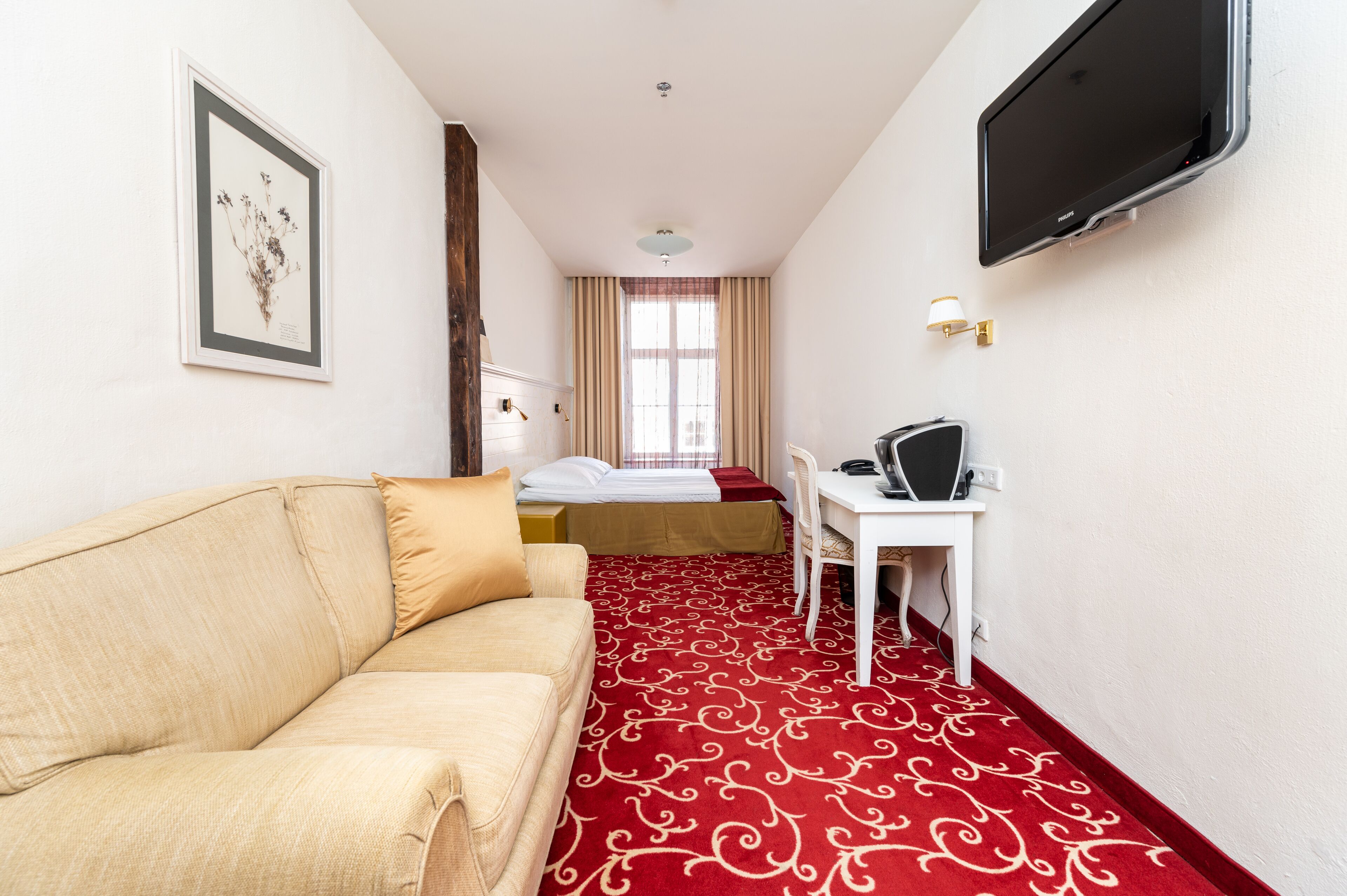 A hotel room with a beige sofa, a bed with a red and white patterned carpet, a desk with a chair, a television mounted on the wall, and a framed picture on the wall.