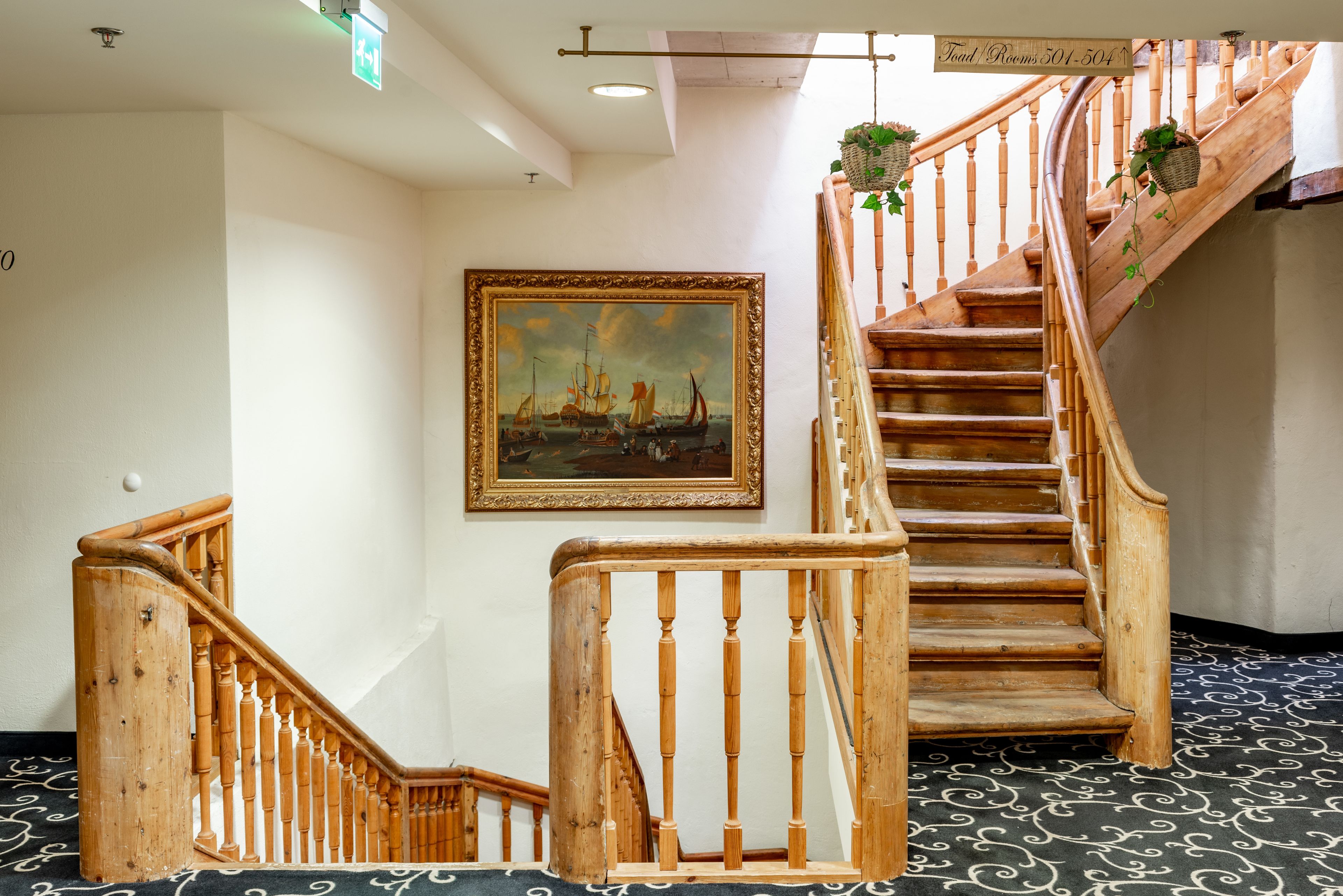 A wooden staircase with a railing, a framed painting on the wall, and a sign indicating room numbers.