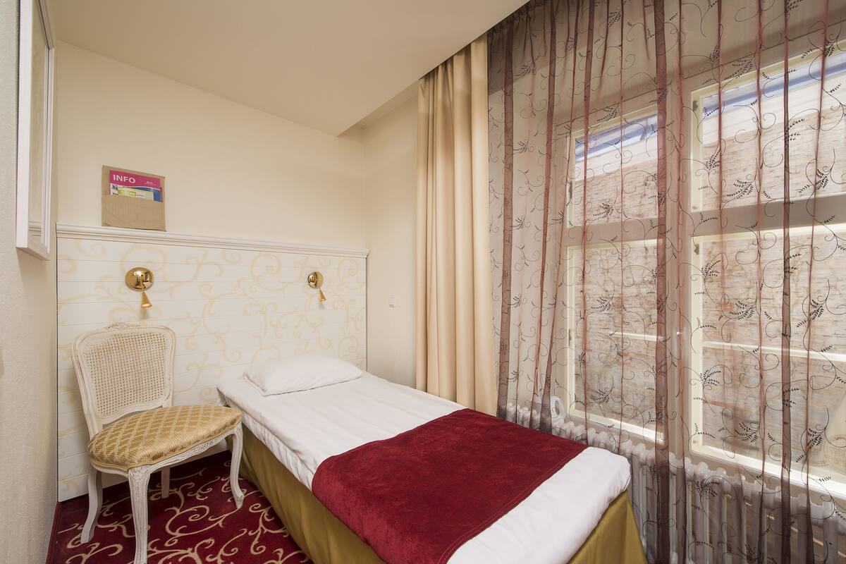 Una cama individual con una manta roja, cabecera blanca, una silla de mimbre y una ventana con cortinas estampadas.