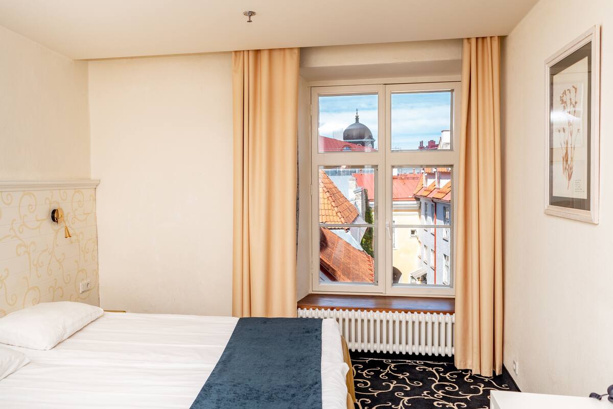 Habitación de hotel con una cama grande, una ventana con cortinas, un radiador y vistas a edificios.