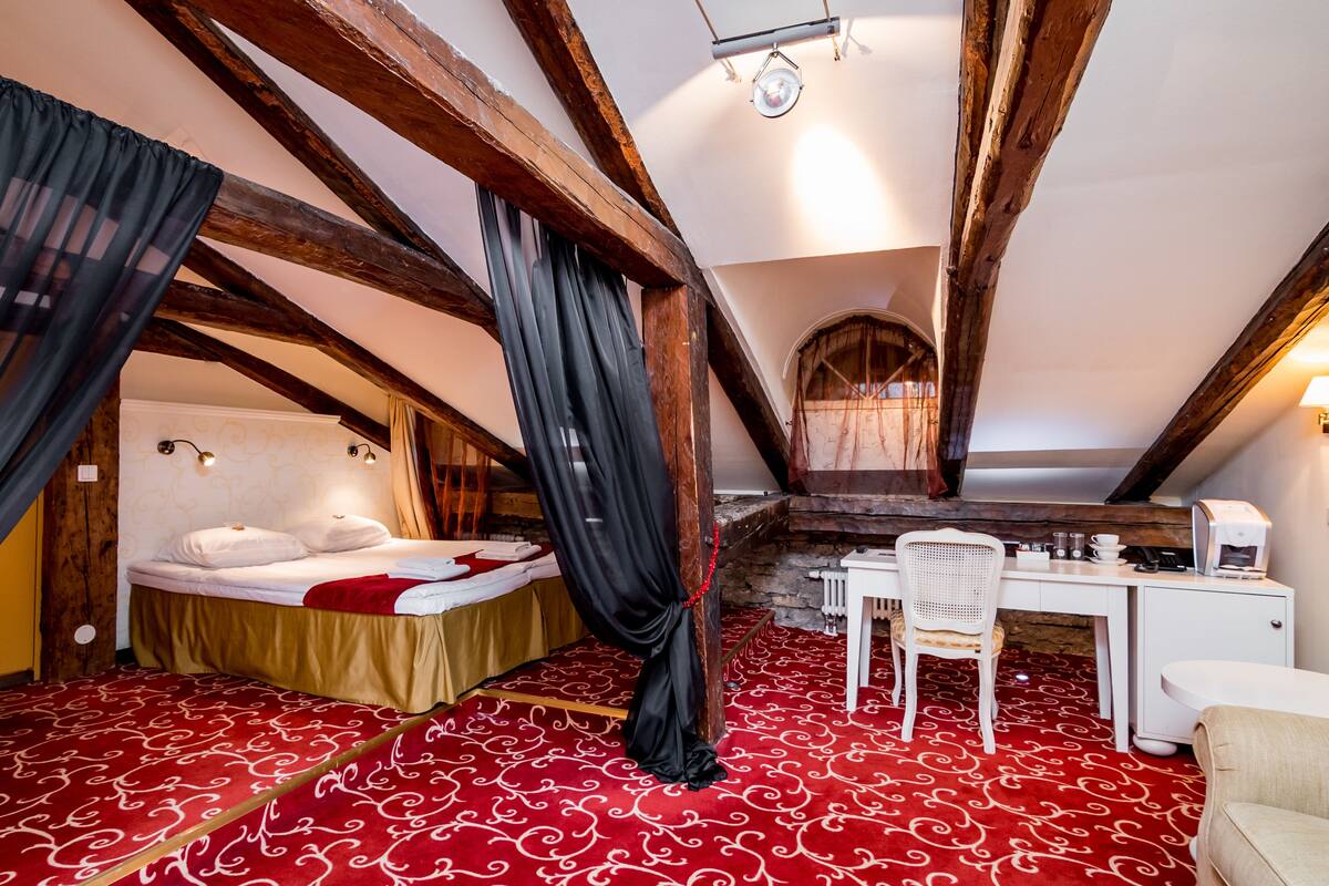 Un dormitorio con una cama, un escritorio y una silla. La habitación tiene vigas de madera al descubierto y una alfombra roja estampada.