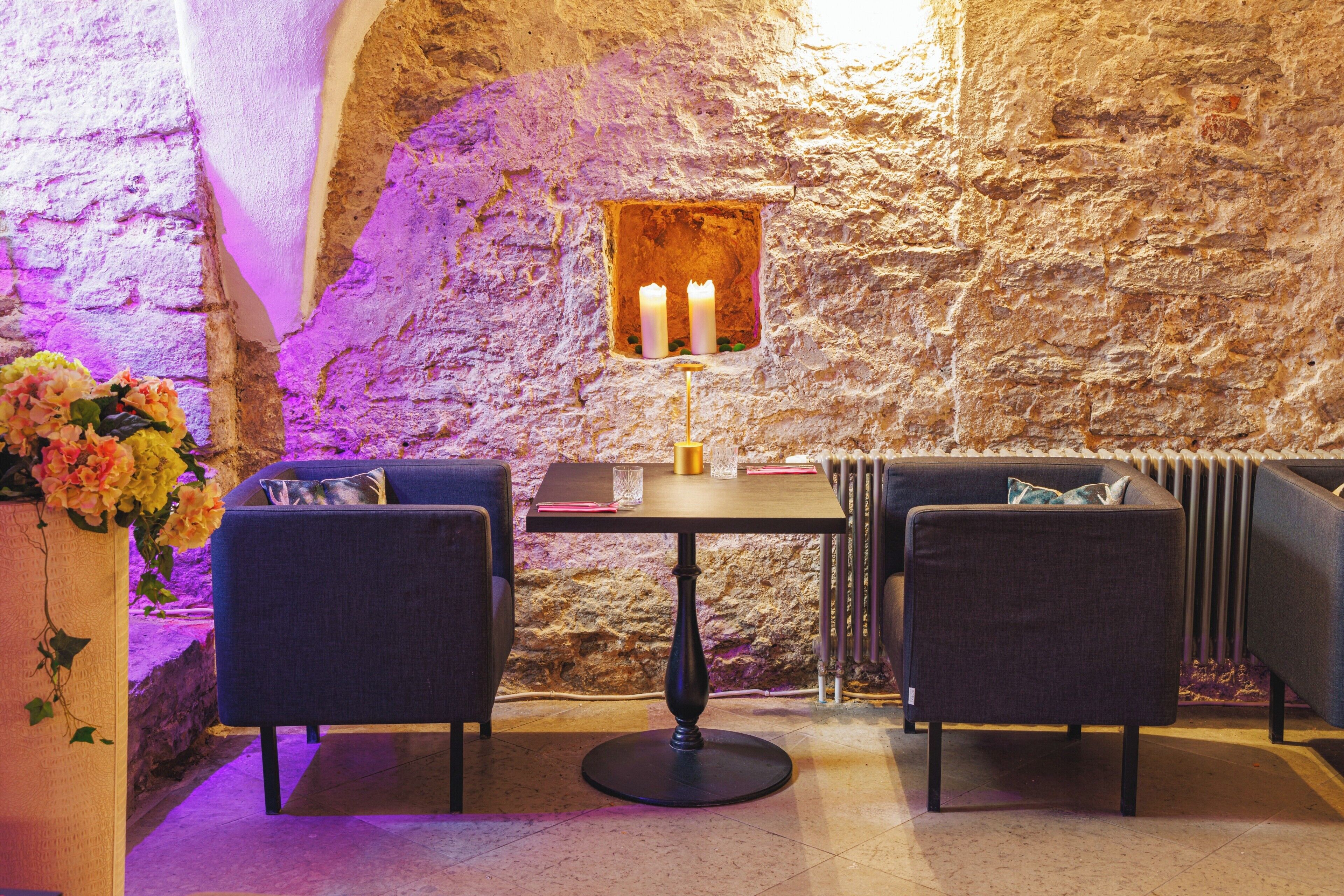 A stone-walled room with a table, two chairs, a vase with flowers, and a lit candle on the wall.