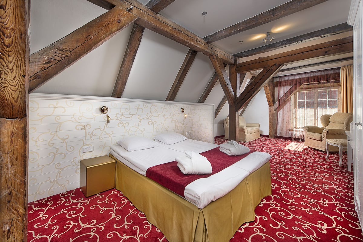 Habitación de hotel con techo alto, vigas de madera a la vista, una cama grande con sábanas blancas, alfombra roja estampada y un área de descanso con dos sillones.