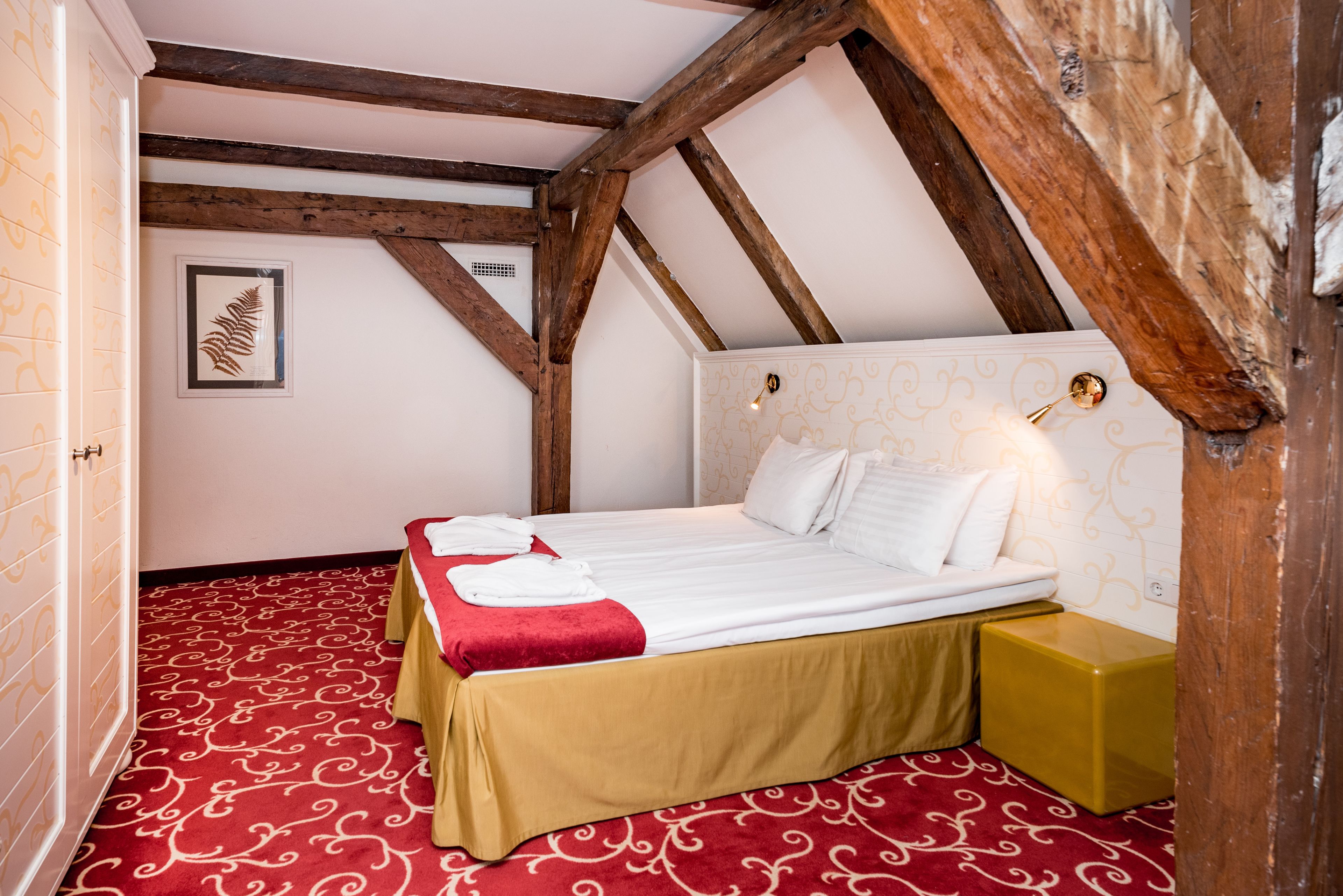 A room with a high ceiling and exposed wooden beams, a bed with white linens, a red patterned carpet, and a small bedside table with a yellow lamp.