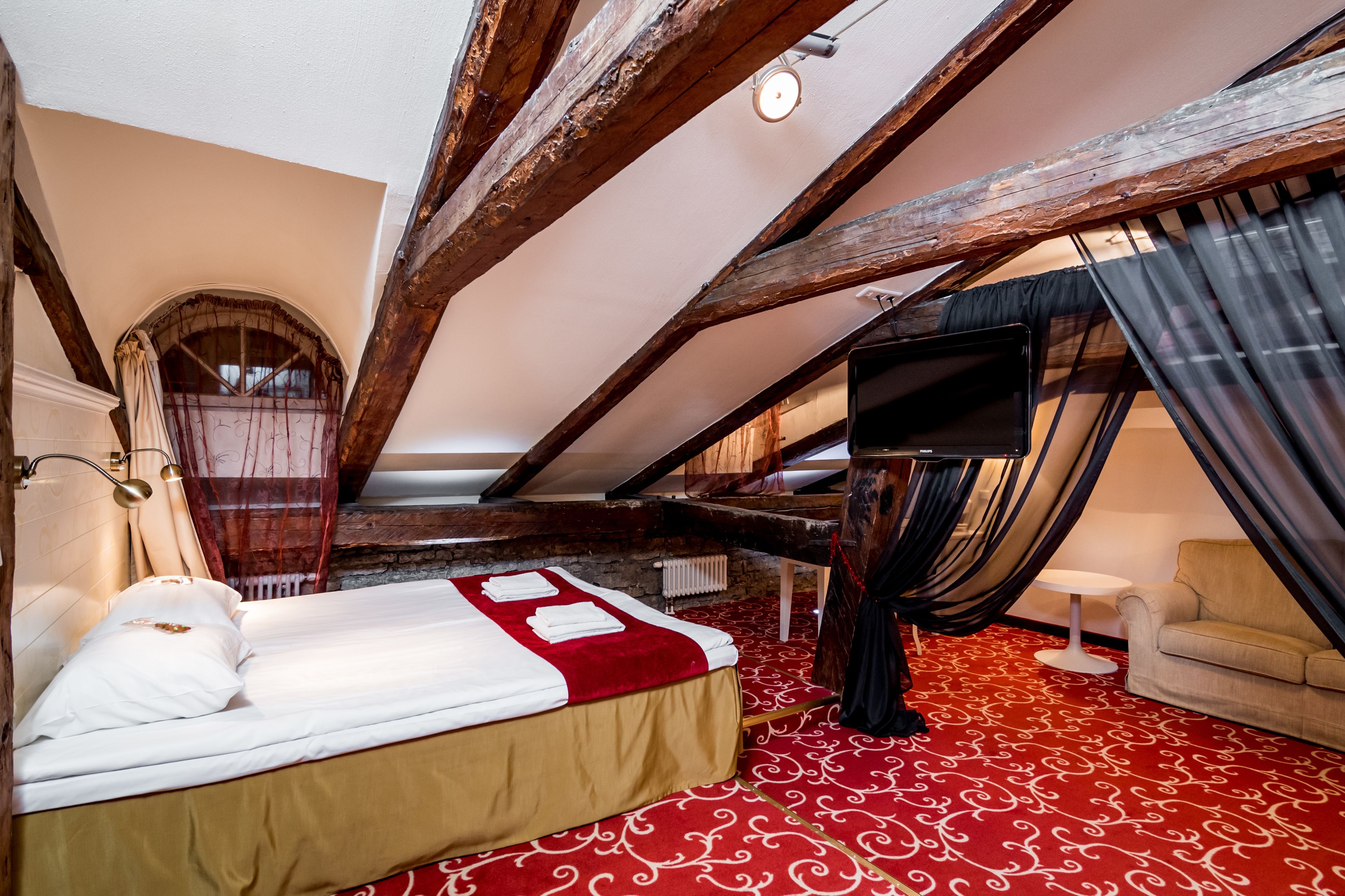 Un dormitorio con una cama, una alfombra roja con diseño, techo de madera, televisor y una pequeña mesa.