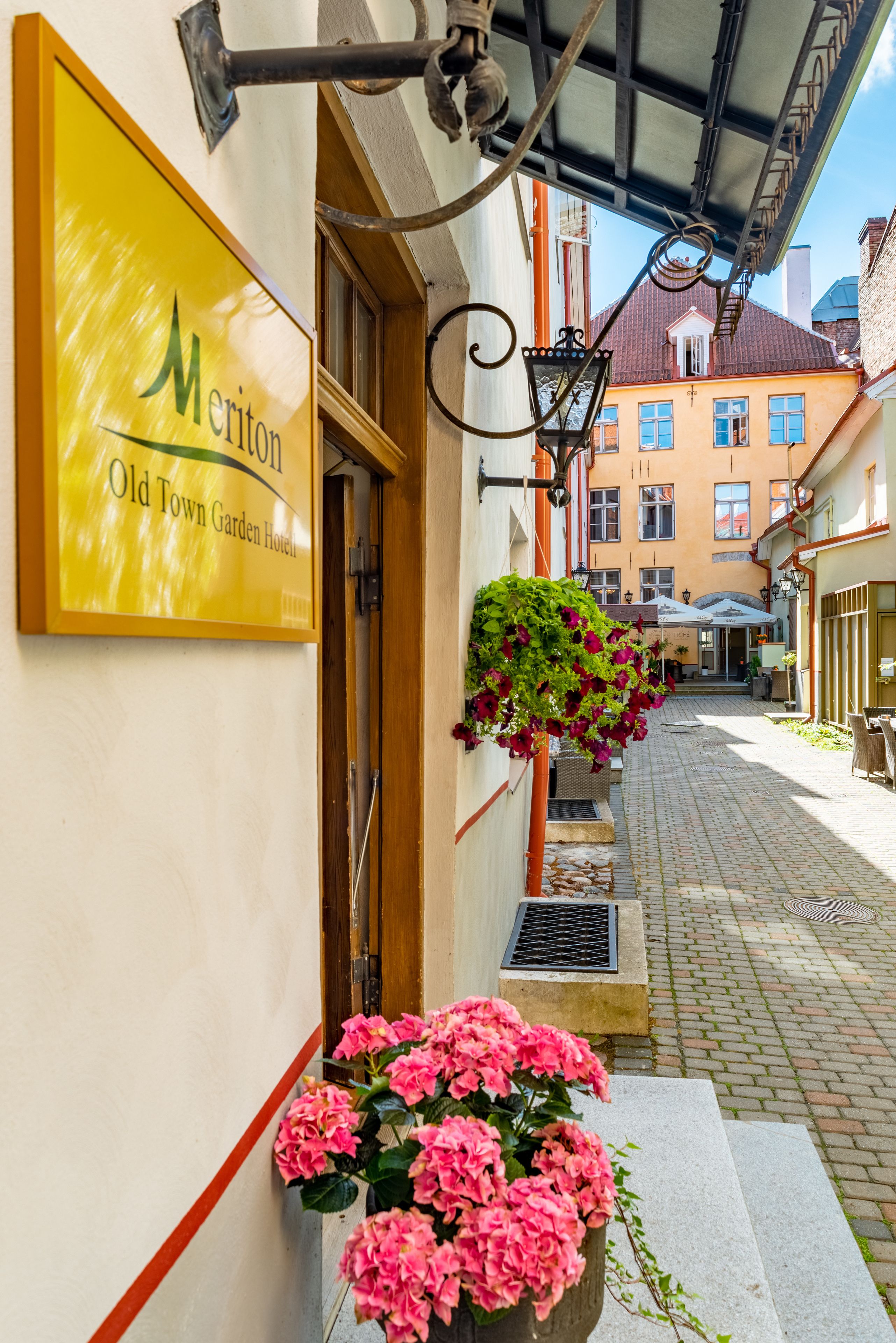 A hotel entrance with a sign for "Meriton Old Town Garden Hotel," a brick pathway, and hanging flower baskets.