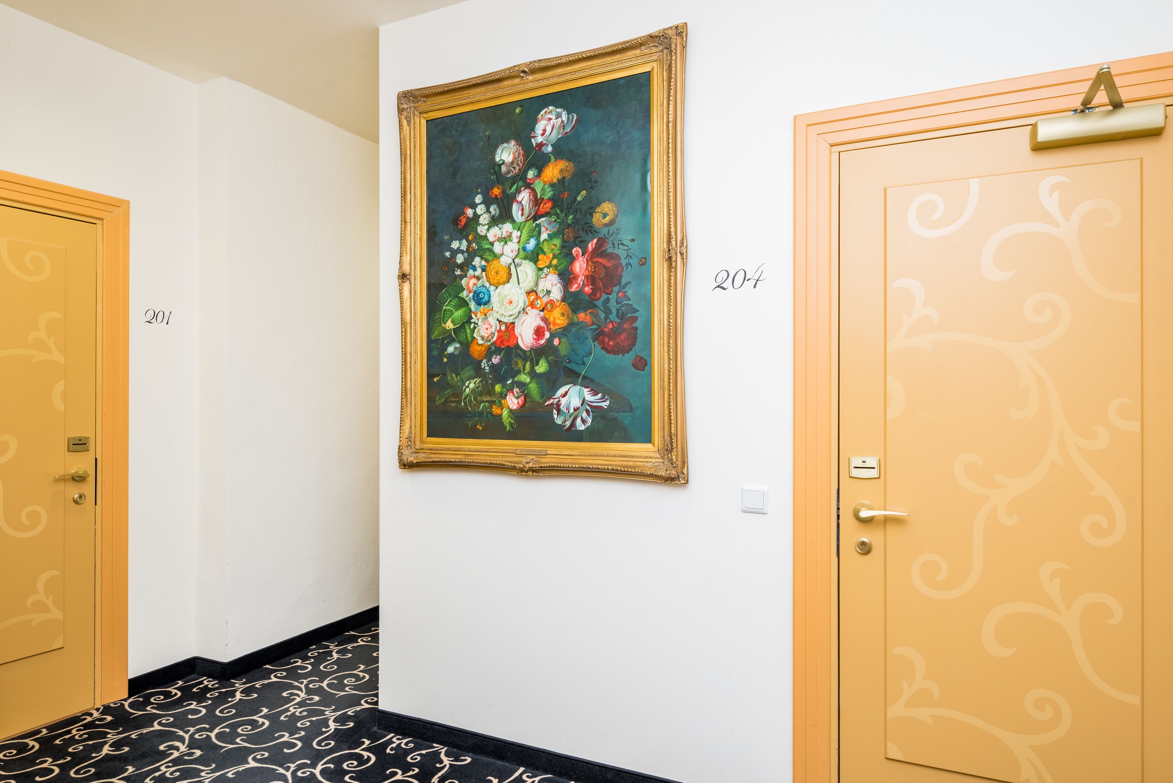 En hall med en inramad blomstermålning på väggen, numrerade dörrar och en mönstrad matta.