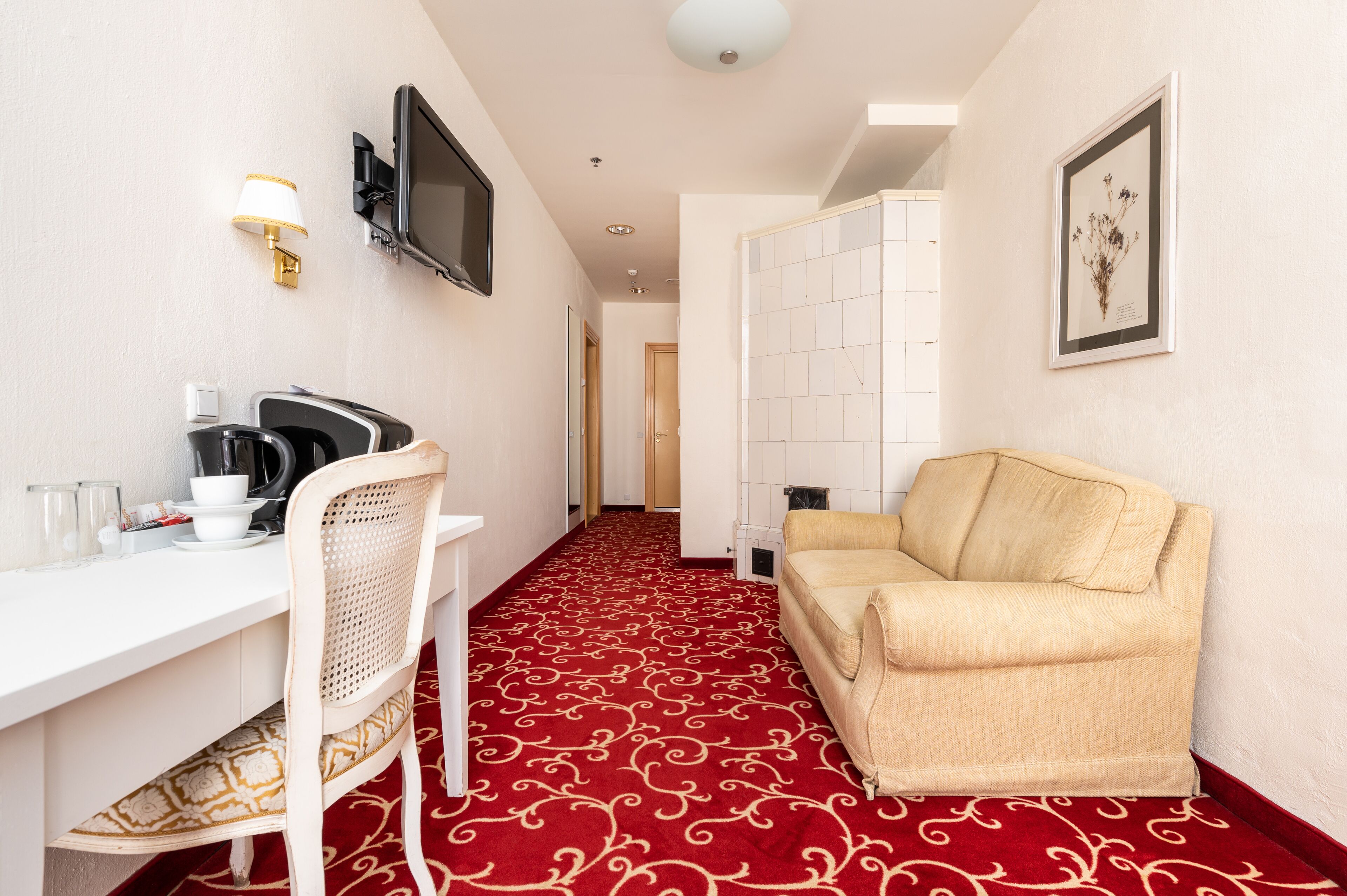 Une chambre d’hôtel avec une moquette rouge, un bureau blanc avec une chaise, un canapé beige, un téléviseur à écran plat fixé au mur et un tableau encadré accroché au mur.