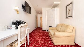 Une chambre d’hôtel avec une moquette rouge, un bureau blanc avec une chaise, un canapé beige, un téléviseur à écran plat fixé au mur et un tableau encadré accroché au mur.
