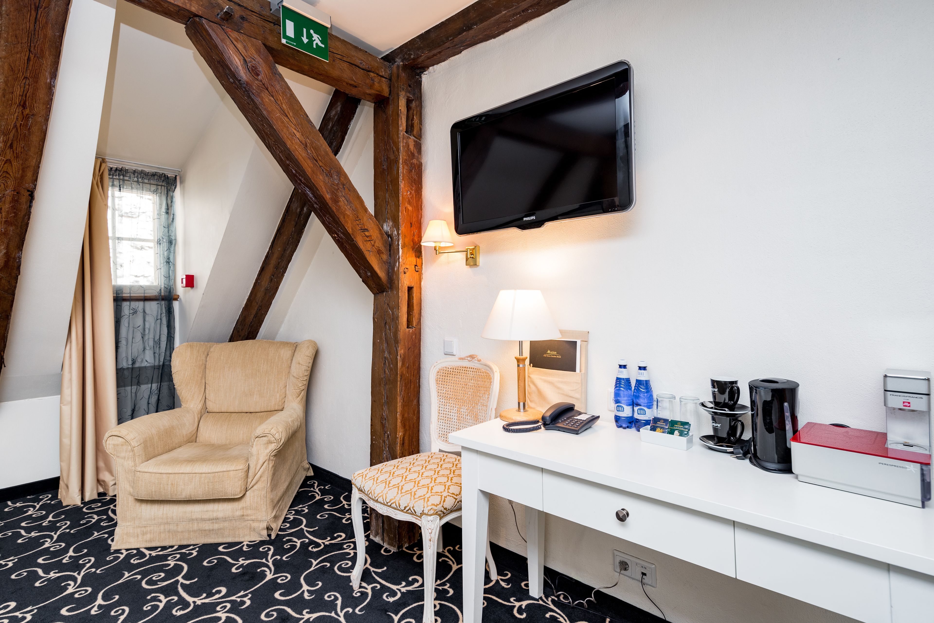 A hotel room with a wooden ceiling, a flat-screen TV, a desk with a lamp, a chair, and a coffee maker.
