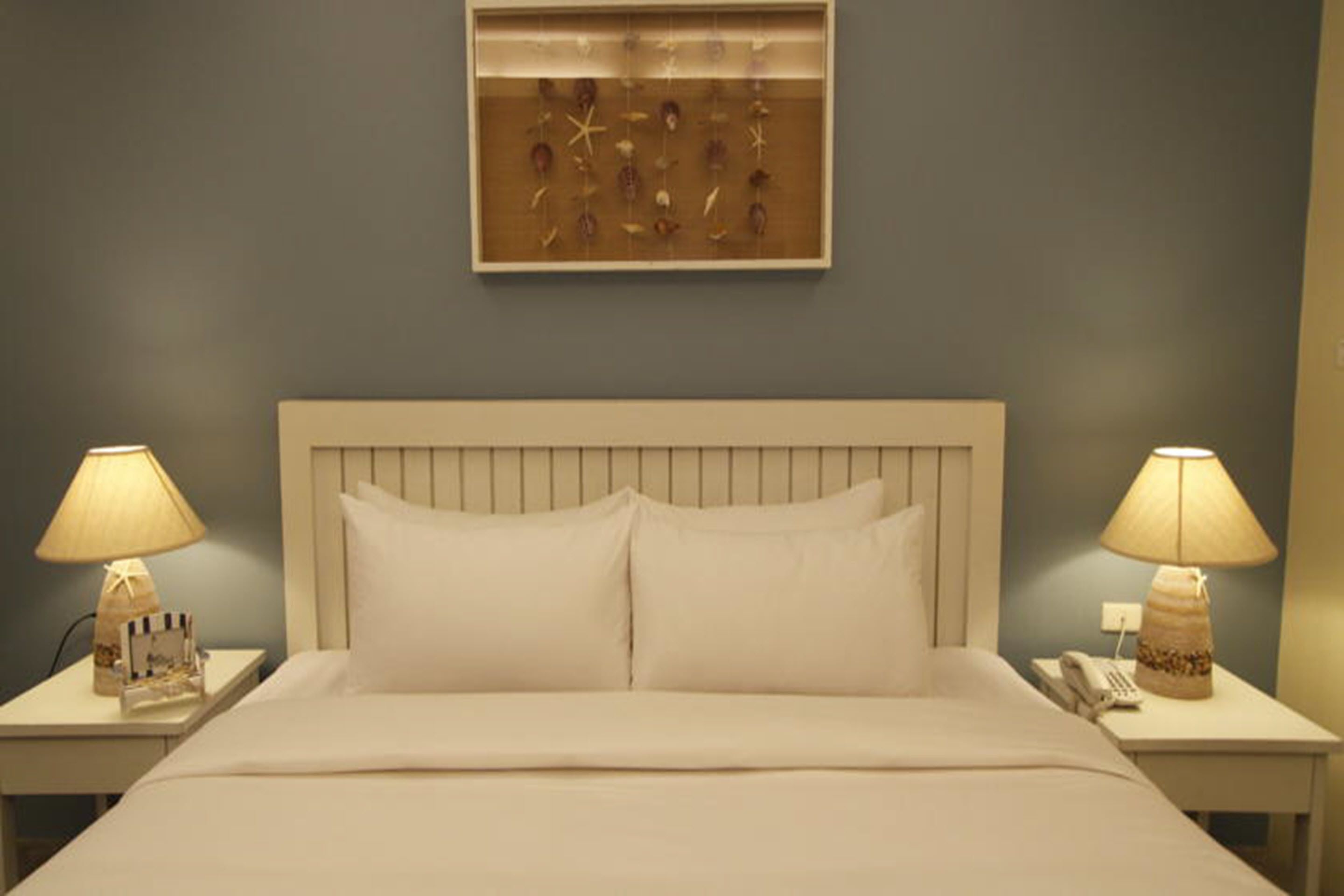 Una habitación de hotel con una cama grande, dos mesitas de noche con lámparas, un cuadro enmarcado sobre la cama y un teléfono instalado en la pared.