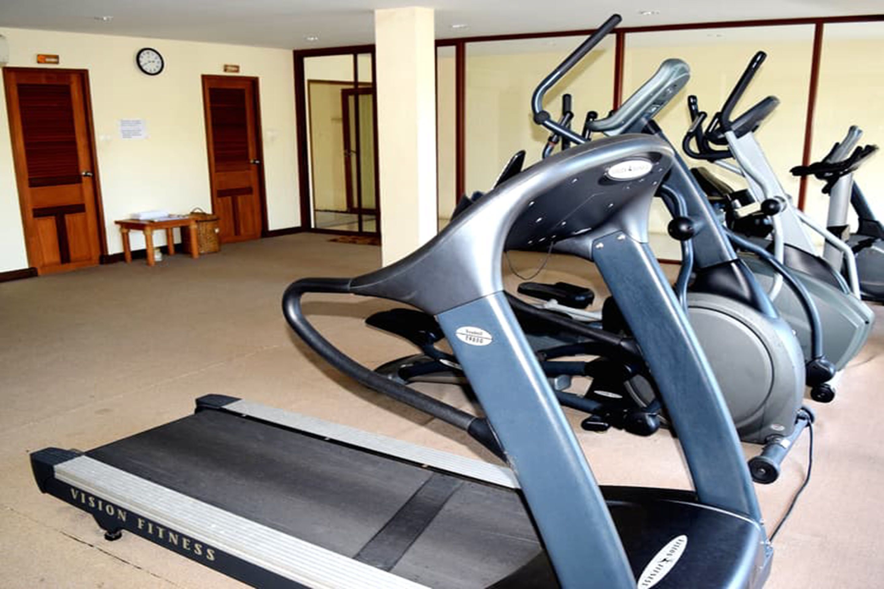 A fitness center with treadmills and elliptical machines. There are wooden doors and a small table with papers on it.