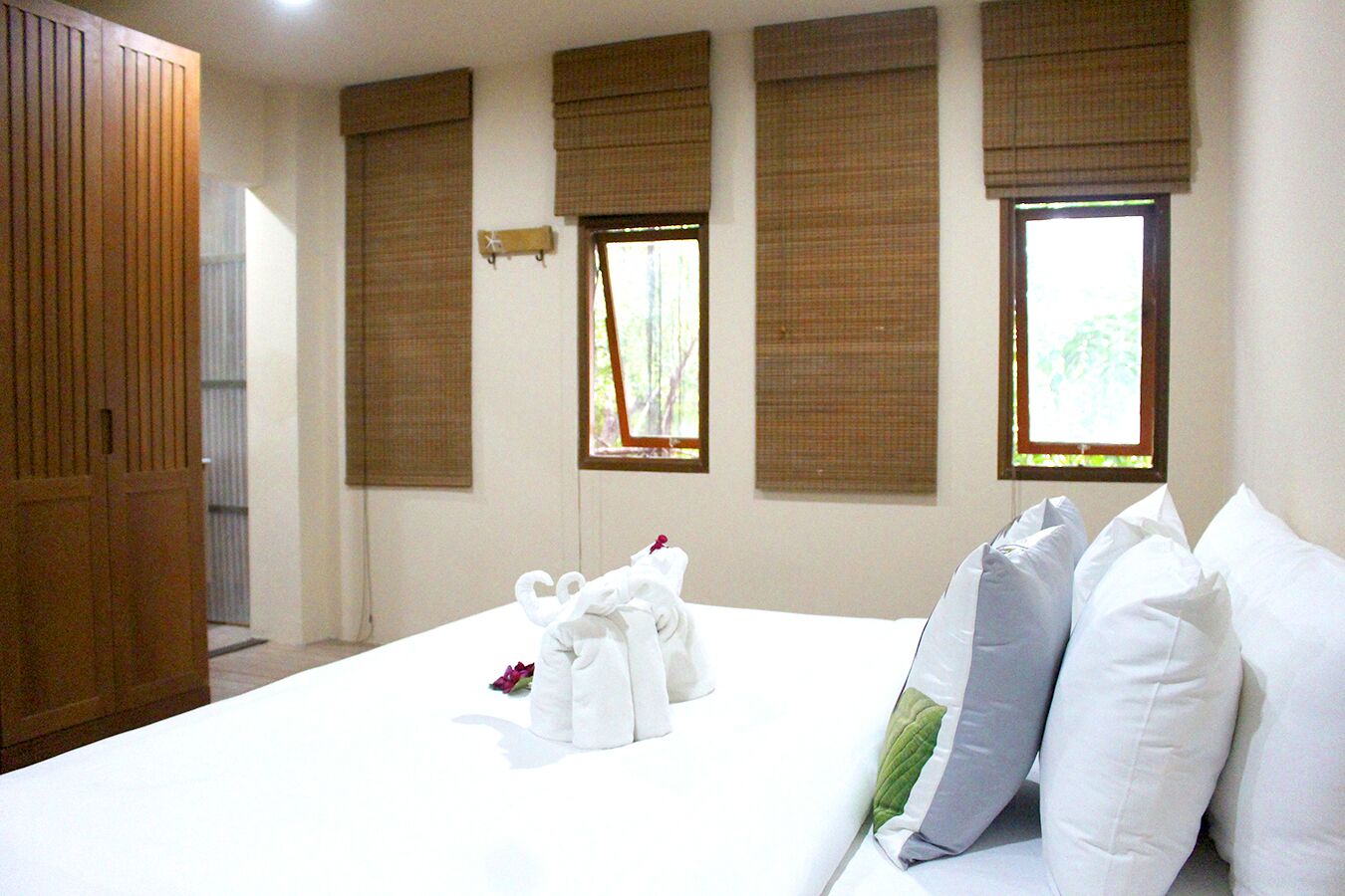 A hotel room with a neatly made bed, a wooden wardrobe, and three windows with brown blinds.