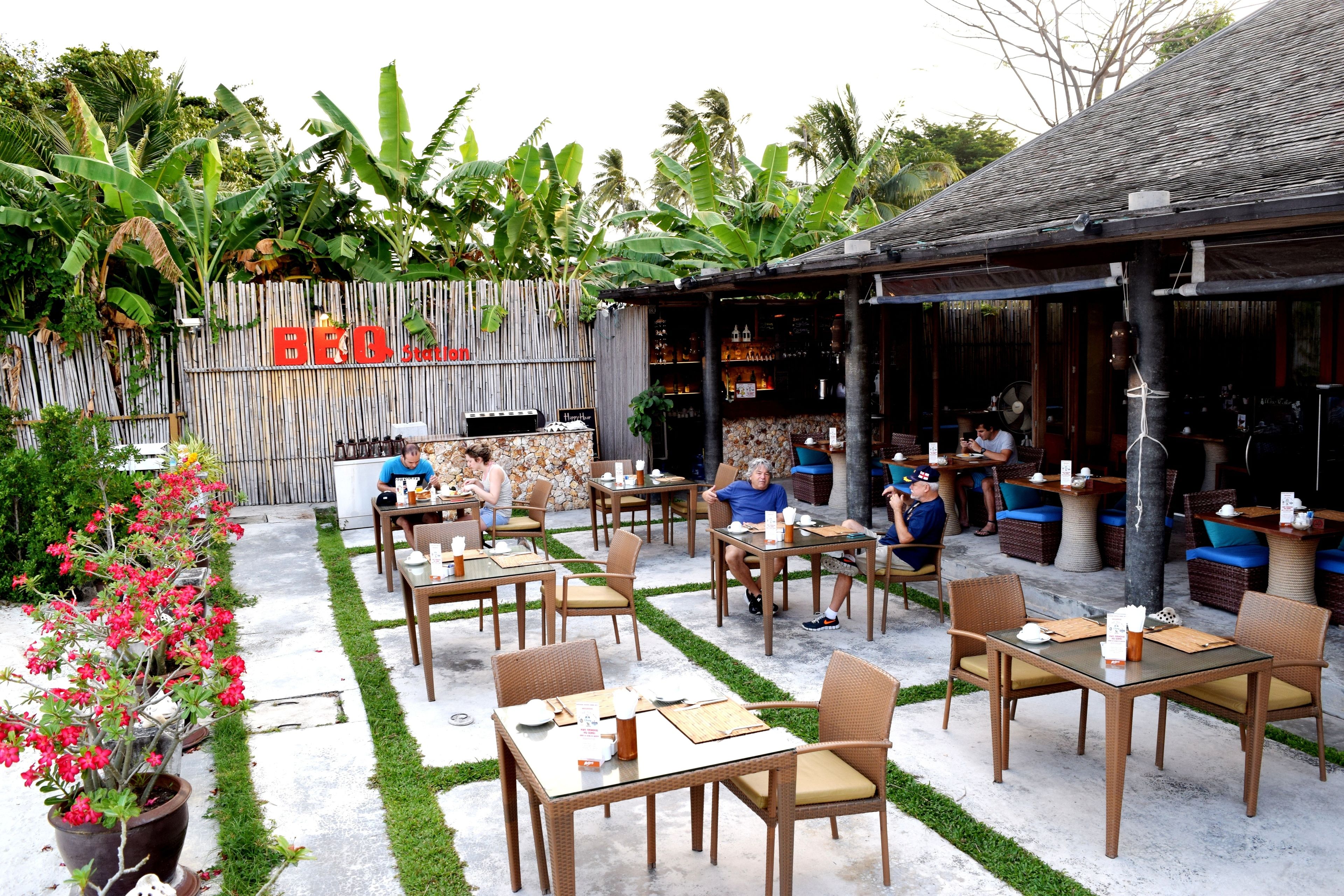 An outdoor restaurant with tables and chairs, surrounded by greenery and a bamboo fence.