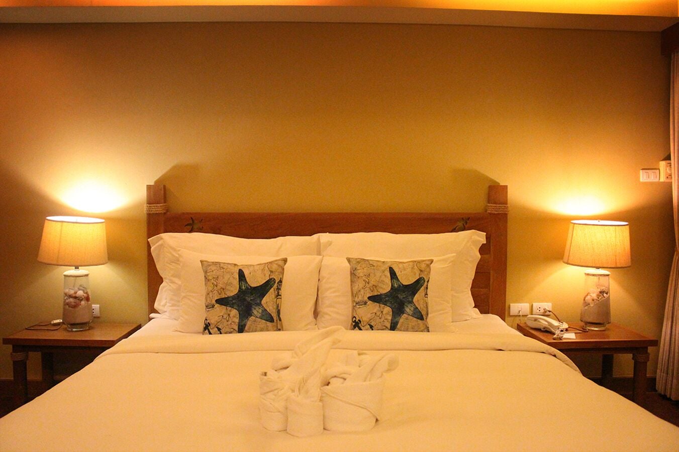 Una habitación de hotel con una cama, dos mesitas de noche con lámparas y dos almohadas con motivos de estrellas de mar.