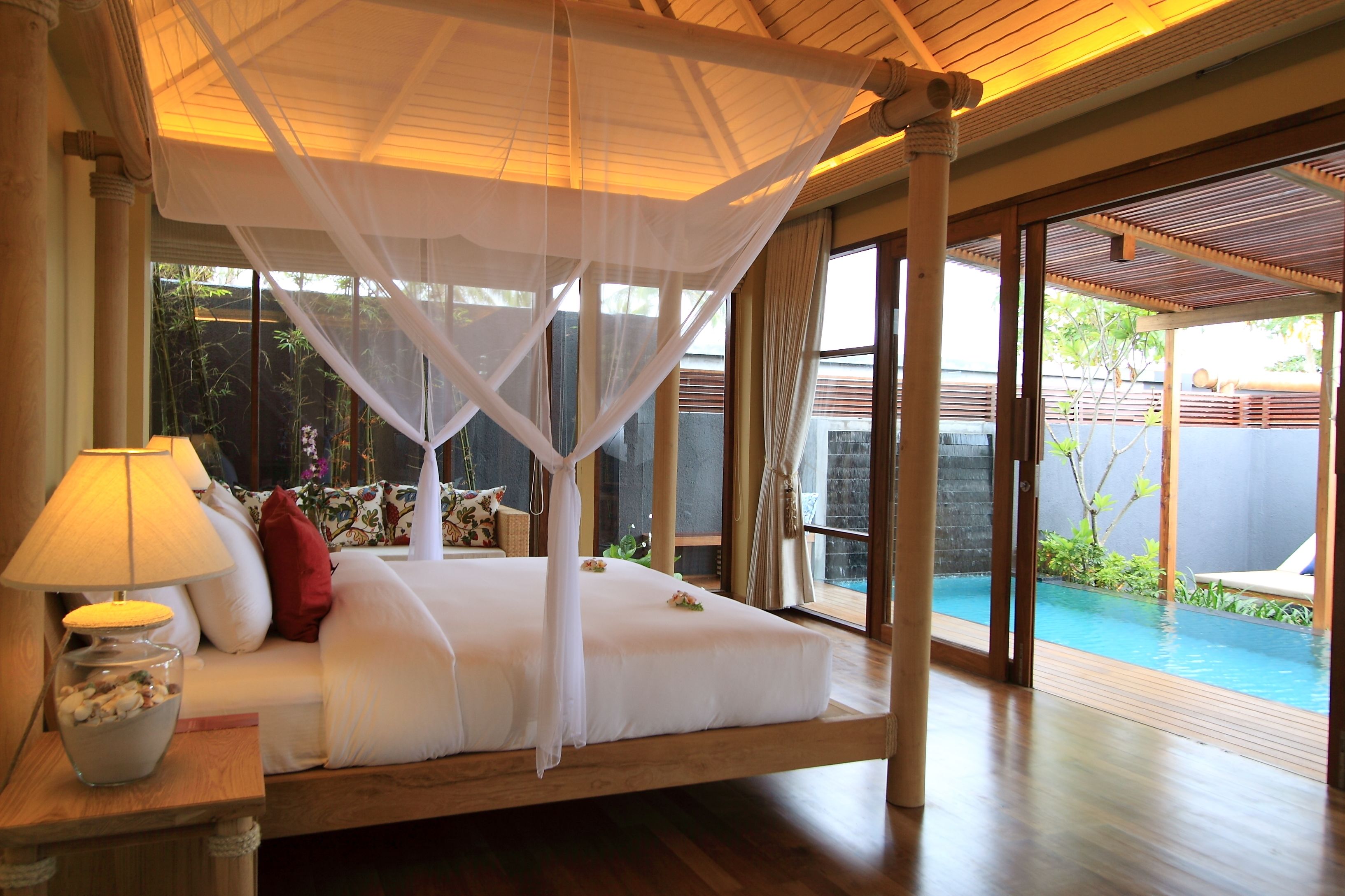 A bedroom with a canopy bed, a bedside table with a lamp, and a view of an outdoor swimming pool.