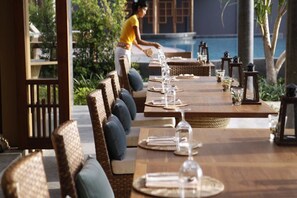 A restaurant with outdoor seating, wooden tables, and chairs, overlooking a pool and greenery.