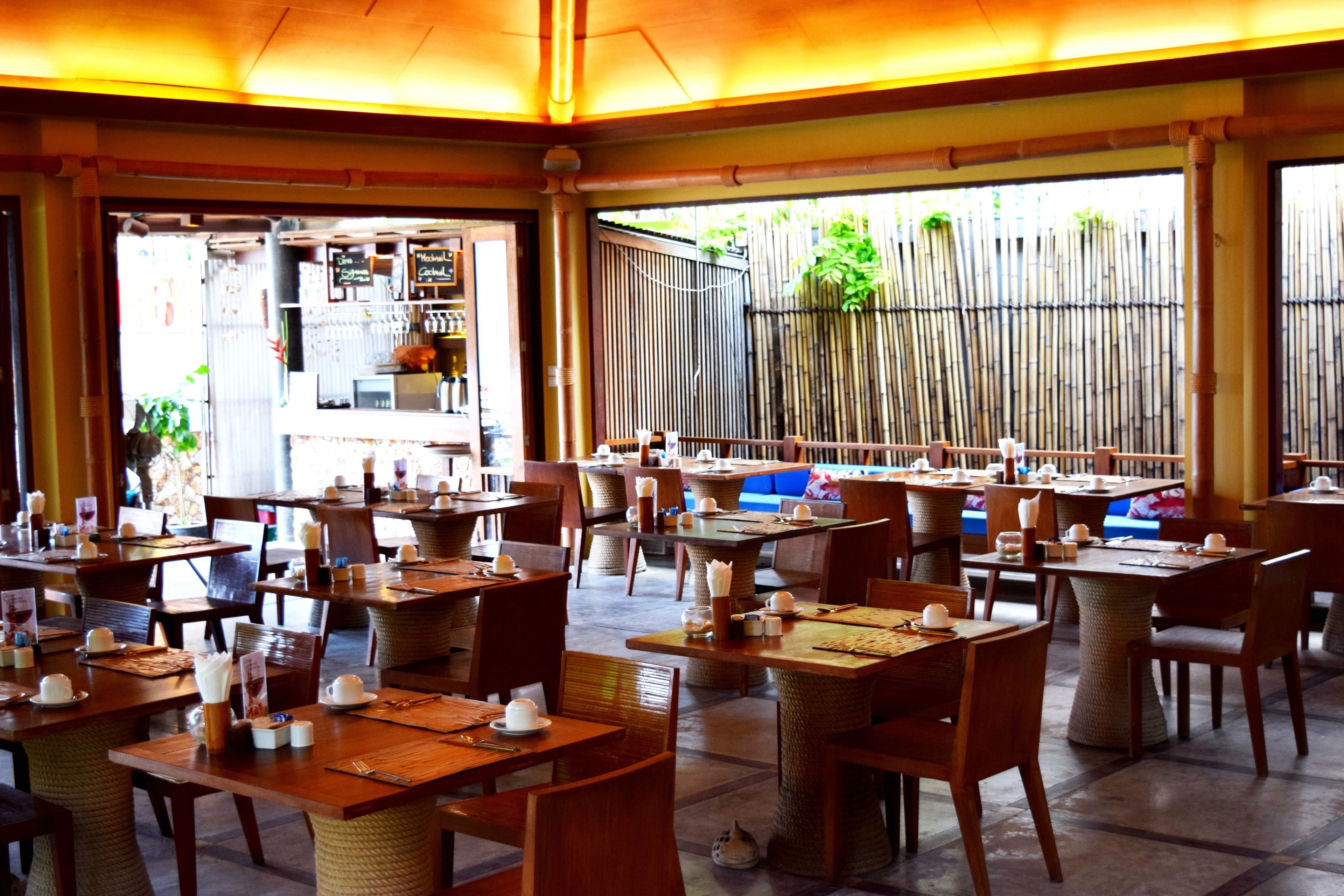 Un restaurante con varias mesas y sillas, paredes de bambú y vistas a zonas verdes al exterior.