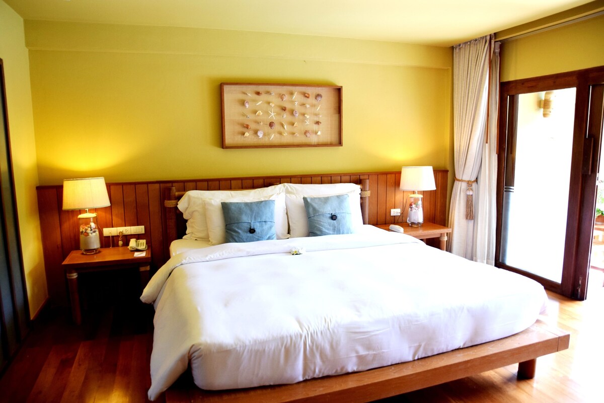 Habitación de hotel con una cama grande, cabecera de madera, dos mesitas de noche con lámparas, una obra de arte colgada en la pared y vistas a través de una ventana con cortinas.