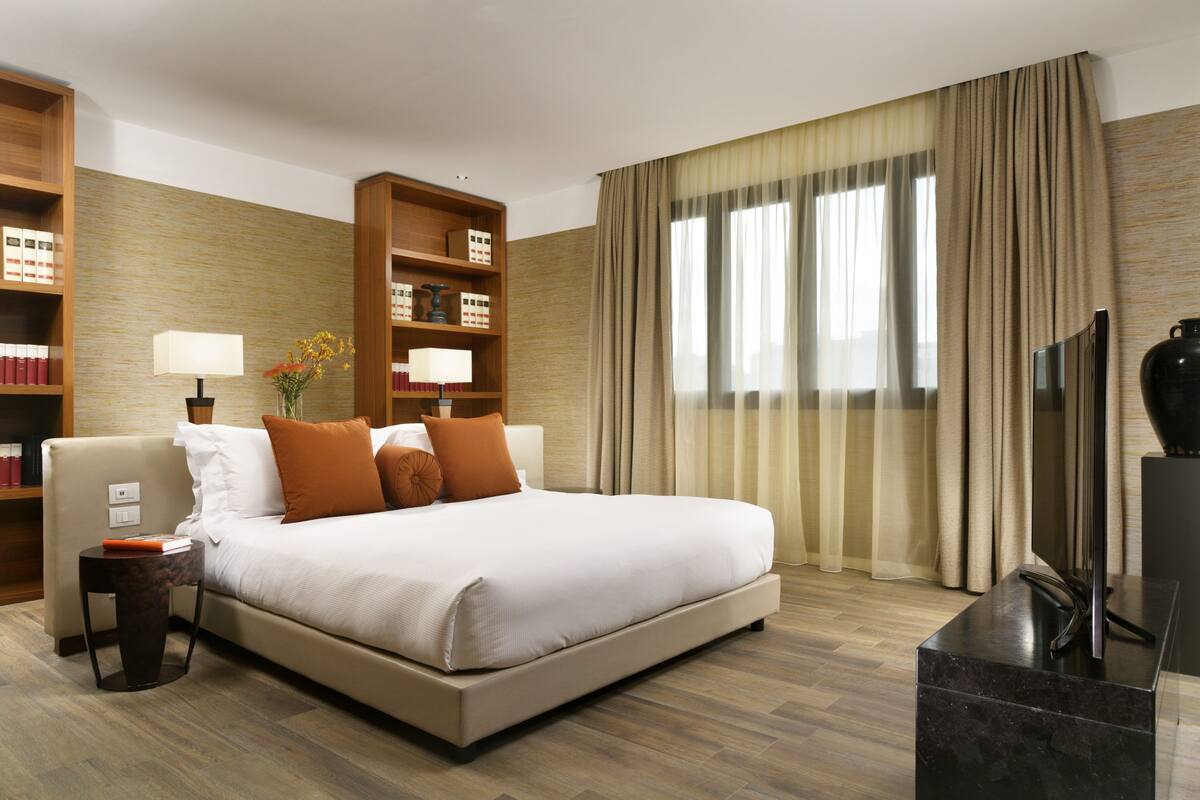 A hotel room with a large bed, a side table, a bookshelf, a TV, and a window with curtains.