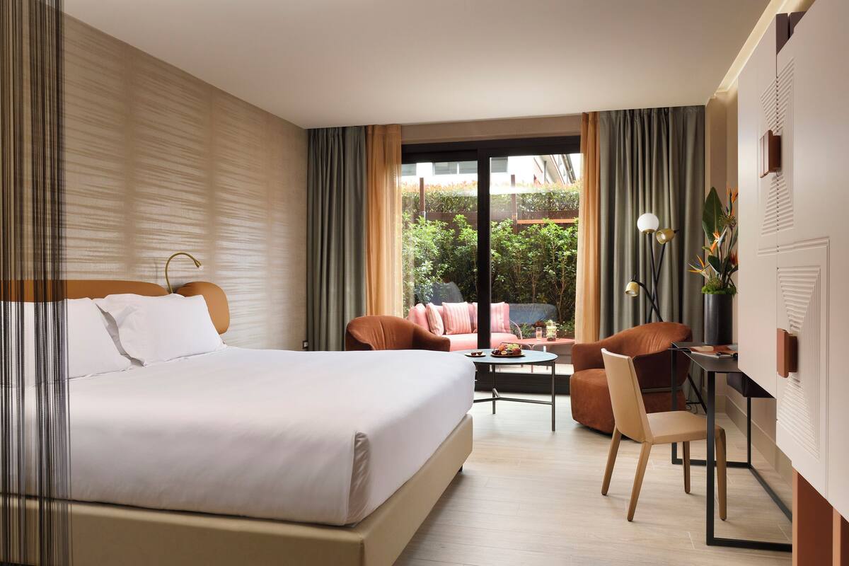 A hotel room with a large bed, a desk, a chair, and a sofa. There is a window with curtains and a view of greenery outside.