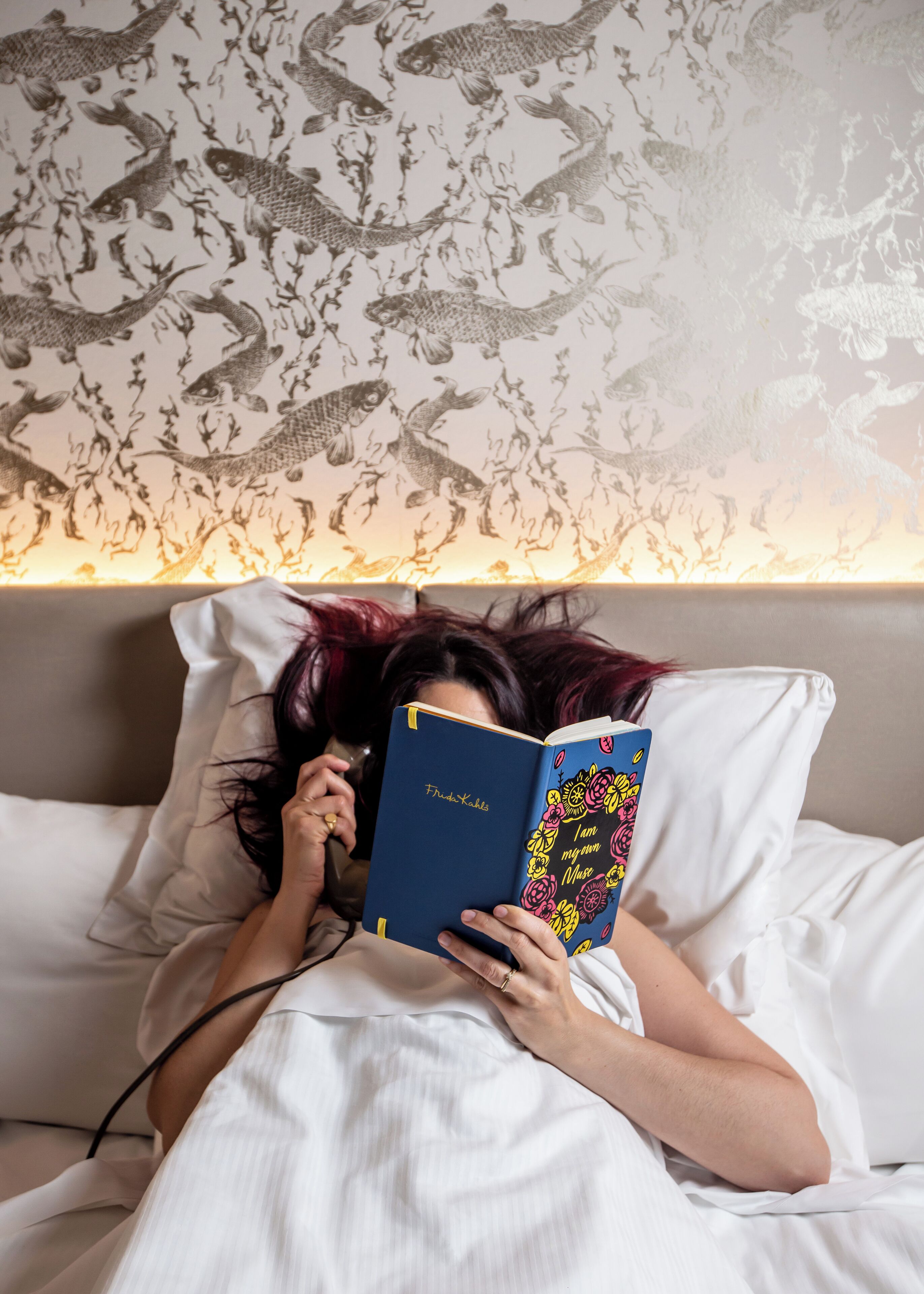 Eine Person liegt in einem Bett, ein Buch mit dem Titel „I am my muse“ sowie eine Tapete mit Fischmotiven sind zu sehen.