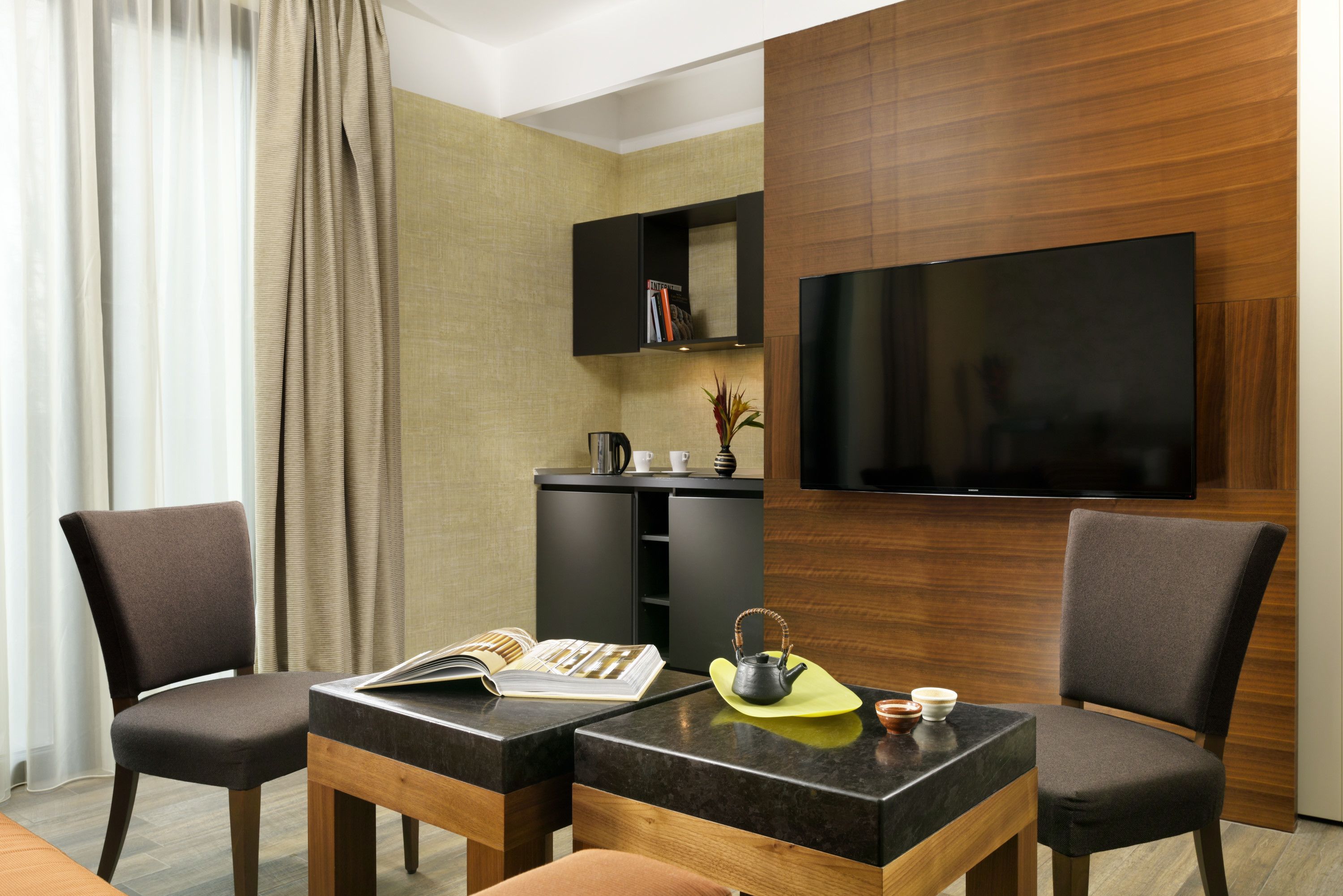 A modern hotel room with a flat-screen TV, a wooden cabinet with books, a small table with a magazine, a teapot, and two chairs.