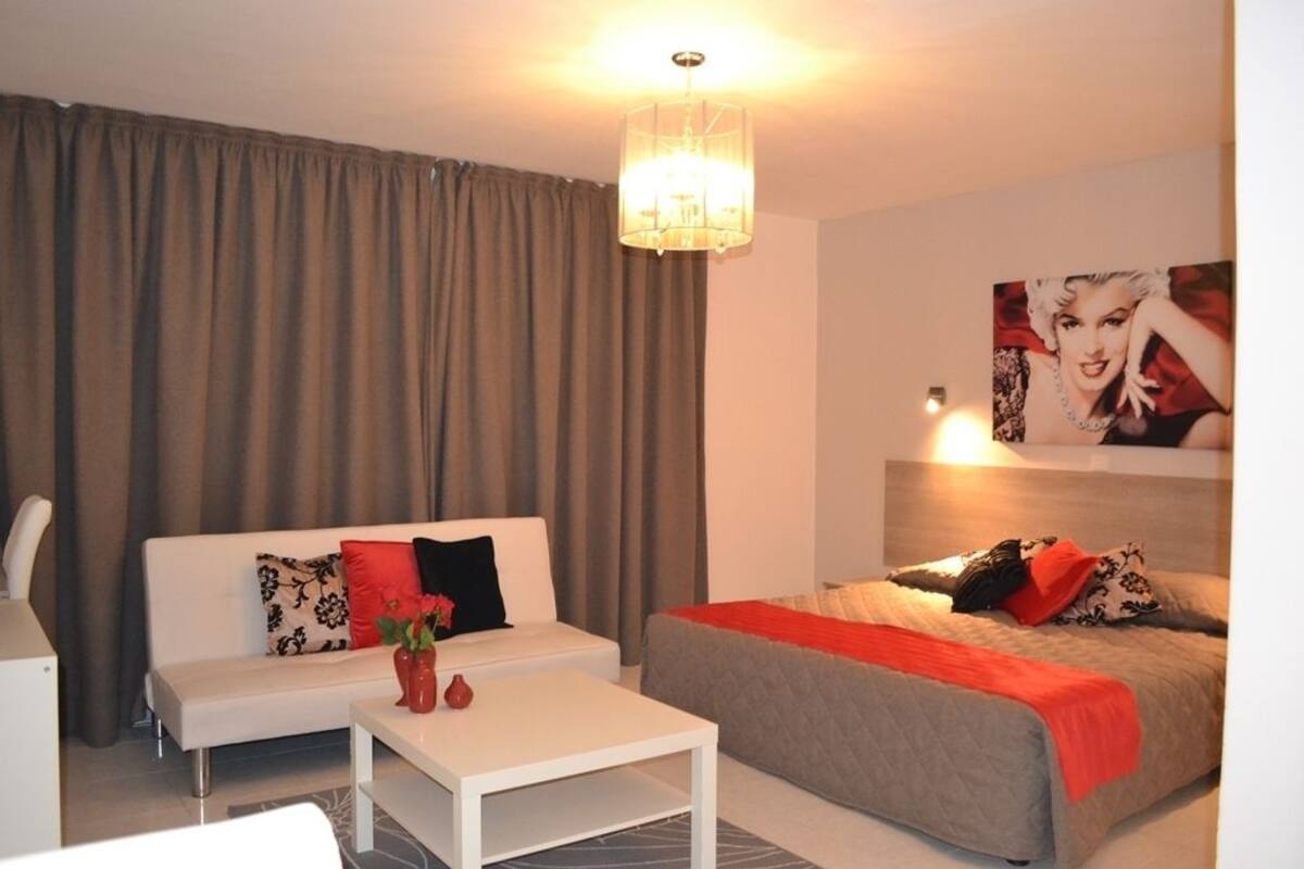 Moderni hotellihuone, jossa on valkoinen sohva, punaisella peitolla varustettu sänky, valkoinen sohvapöytä ja kattokruunu.