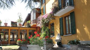 A yellow building with green shutters and a balcony with purple flowers. There are potted plants and a stone bench in the foreground. - Hotel Centrale (Bellagio)