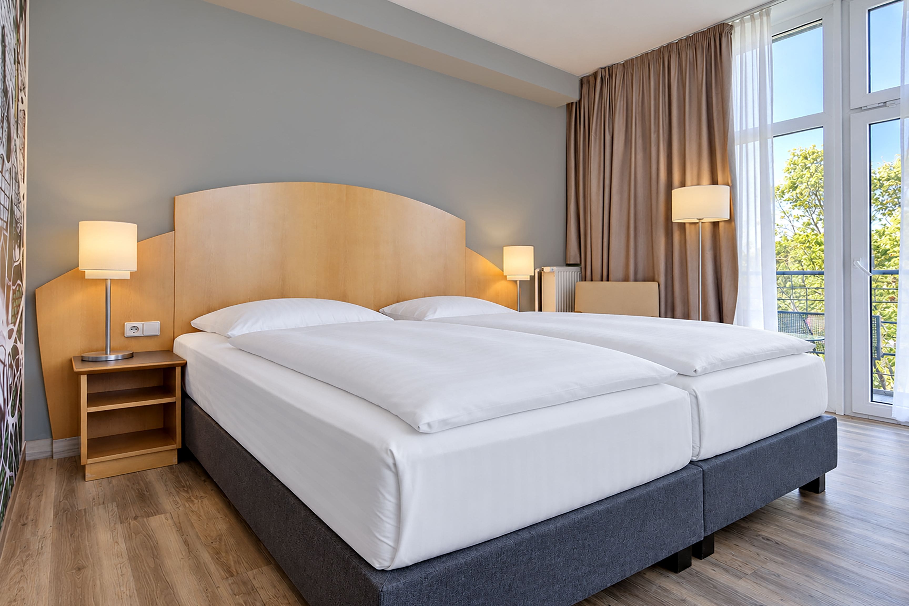 A double bed with a wooden headboard, two bedside tables with lamps, a view of greenery outside, and a large window with curtains.