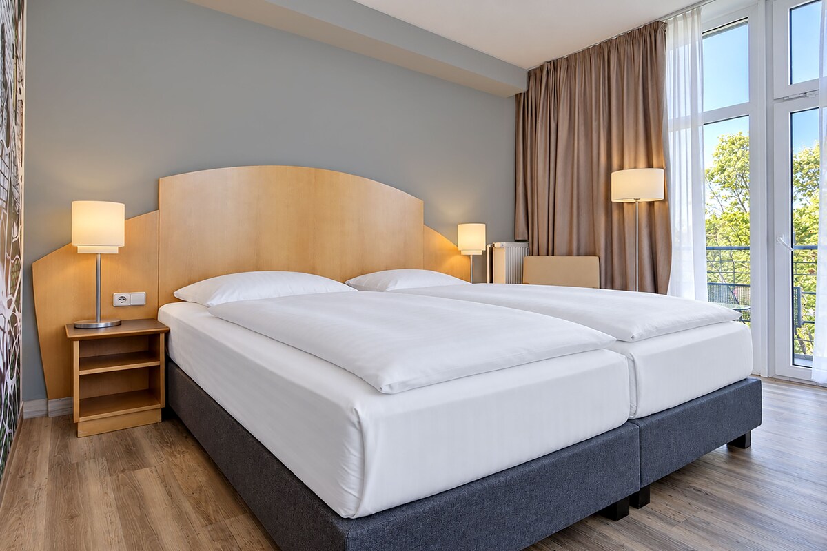 A double bed with a wooden headboard, two bedside tables with lamps, a view of greenery outside, and a large window with curtains.