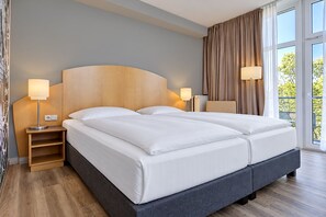 A double bed with a wooden headboard, two bedside tables with lamps, a view of greenery outside, and a large window with curtains. - Seminaris Sunday Hotel Potsdam Babelsberg (Potsdam)