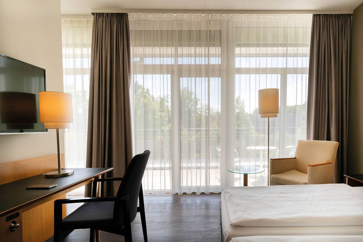 A hotel room with a bed, a desk with a lamp, a chair, a TV, and large windows with curtains.