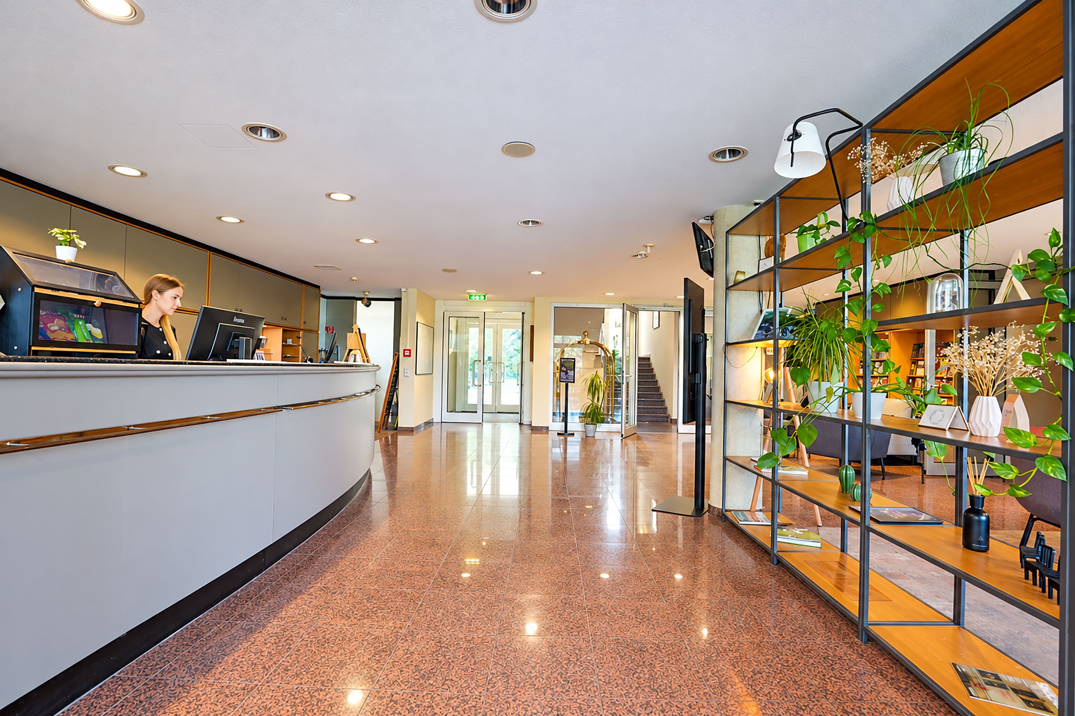 Un espace d’accueil d’hôtel comprenant un comptoir, un bureau d’accueil, un escalier et une étagère ornée de plantes.