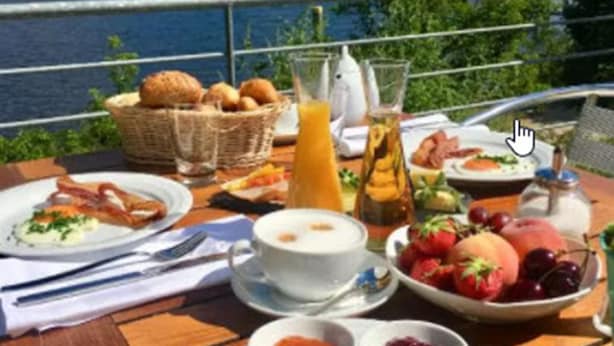 Une table de petit-déjeuner avec divers plats, notamment des œufs, des toasts et des fruits, le tout disposé devant un paysage pittoresque au bord du lac.