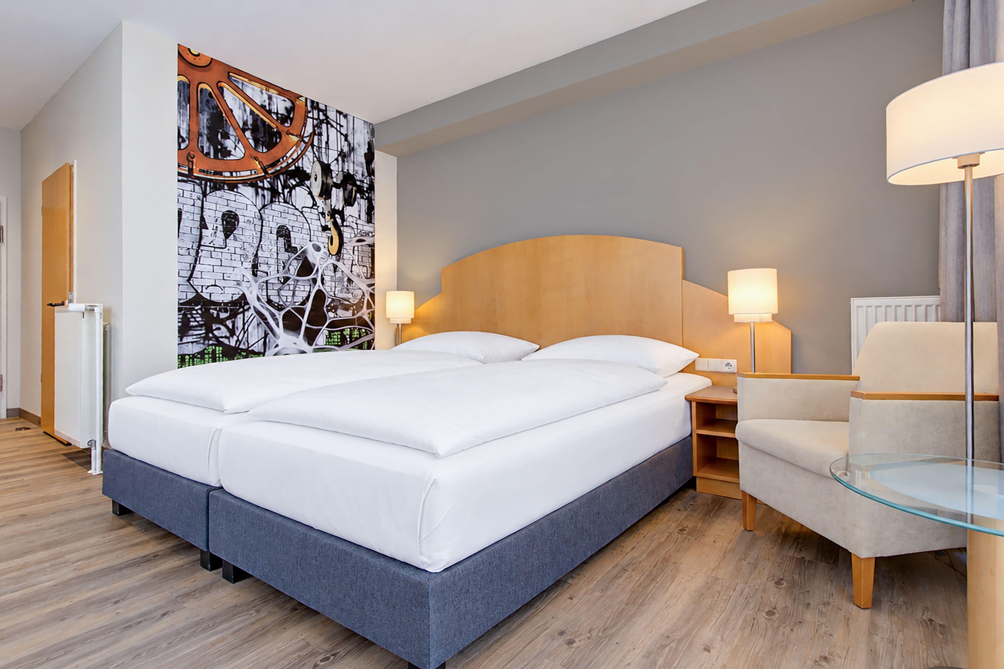 A modern hotel room with a large bed, two bedside lamps, a glass table, and a wall with abstract art.