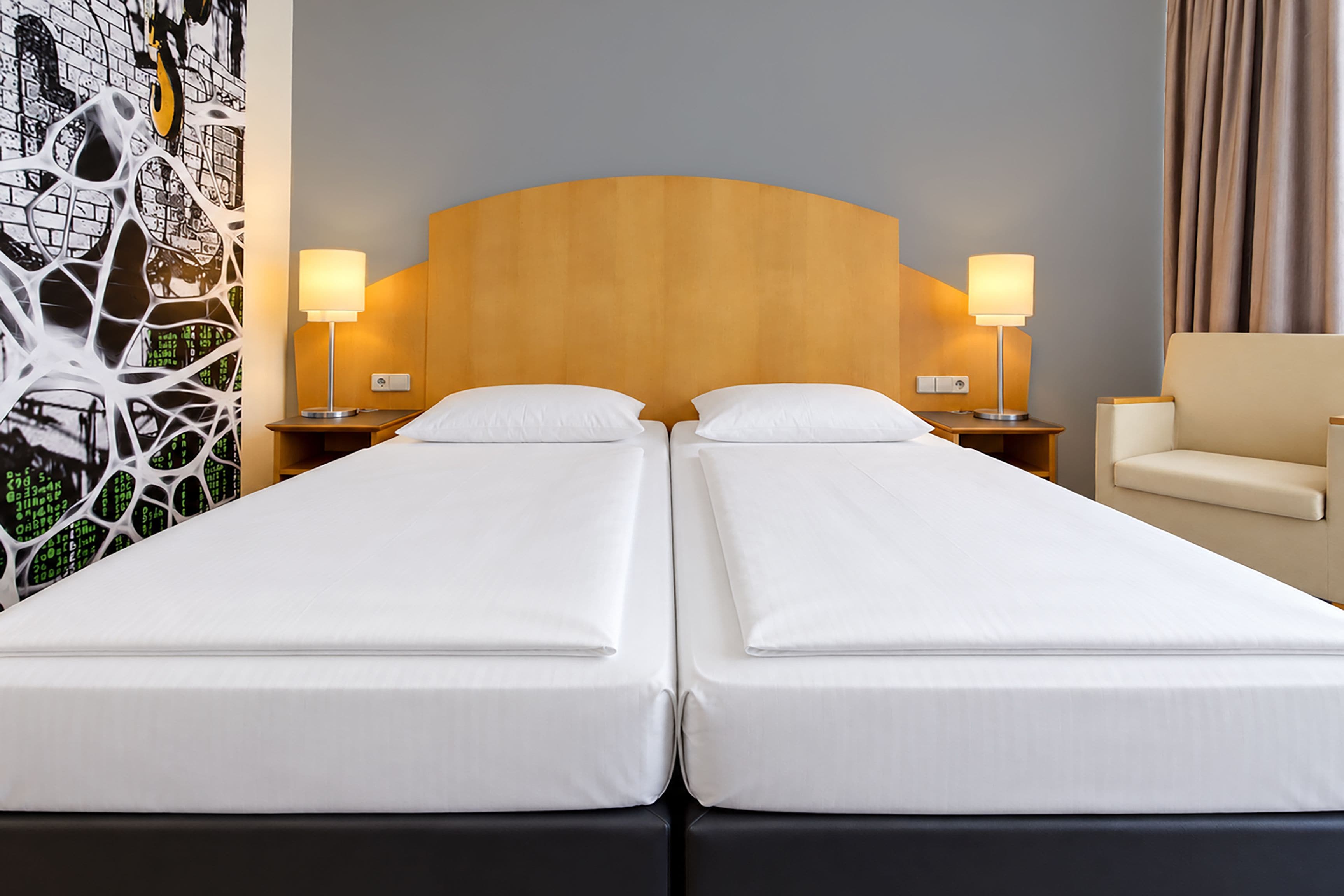 A hotel room with two single beds, a wooden headboard, bedside lamps, a chair, and a wall with a digital art display.